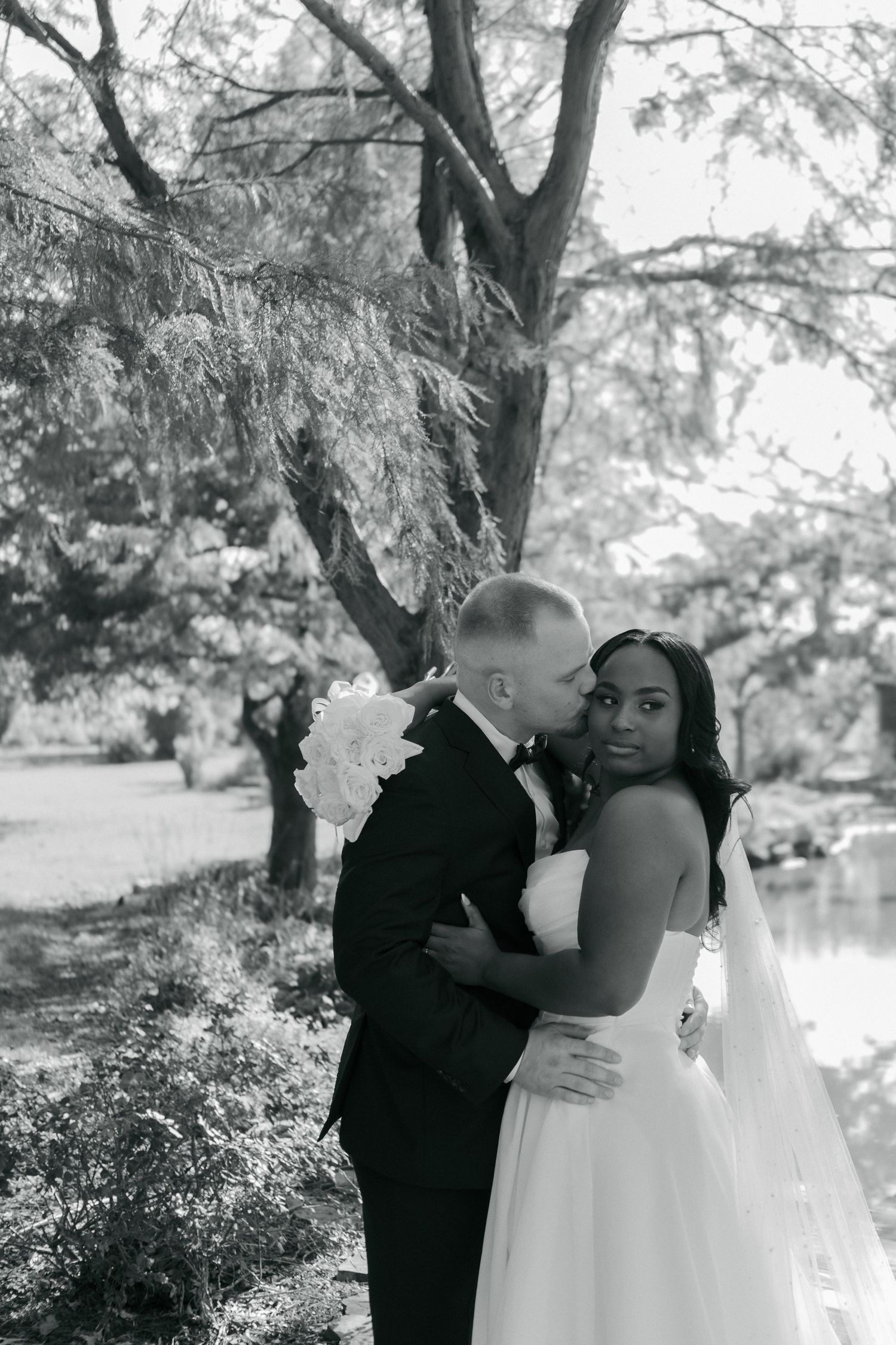 Black and white portrait of groom kissing bride on cheek as she smiles near garden pond.