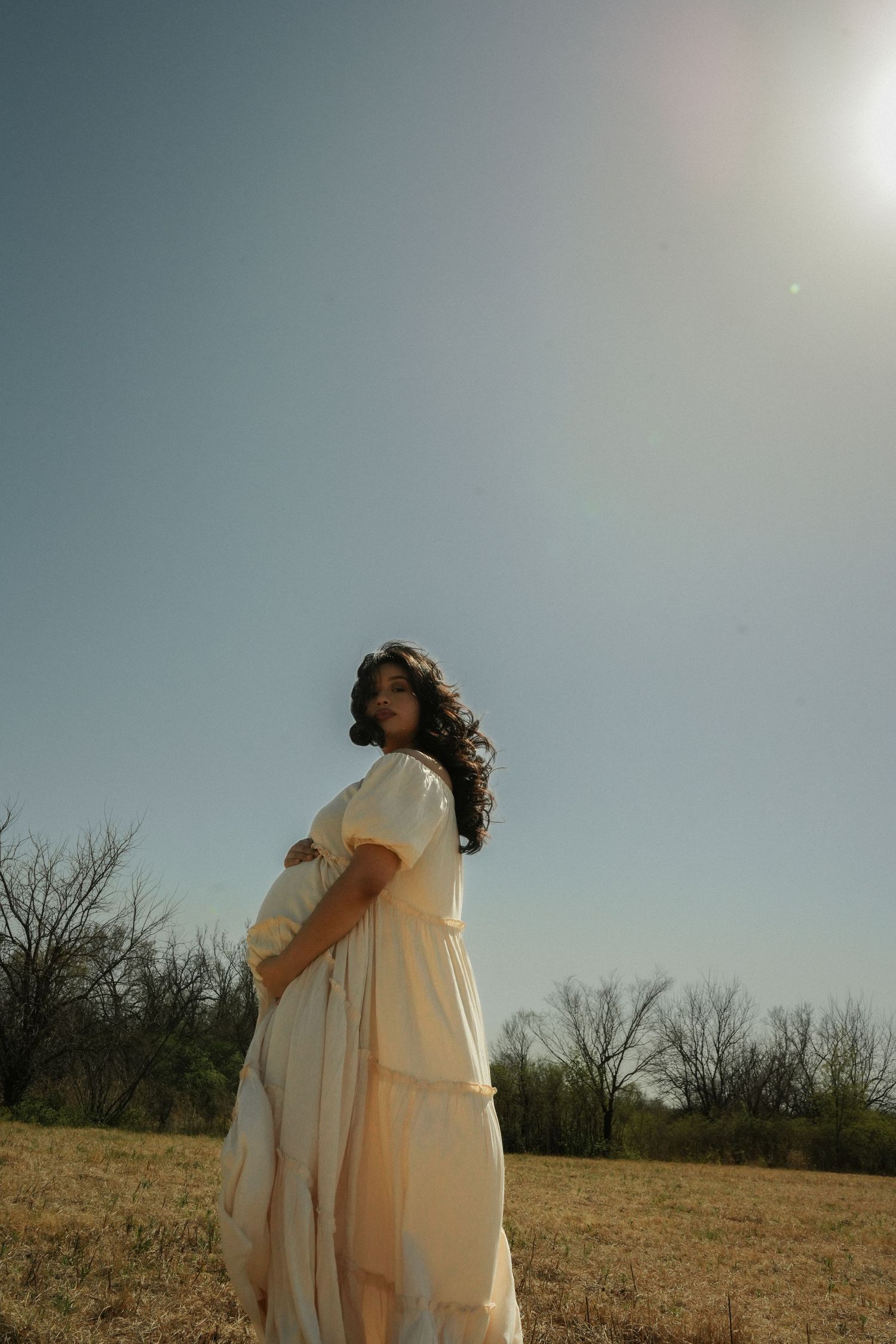 Pregnant woman in flowing white dress holds baby bump in sunny field with bare trees in background.