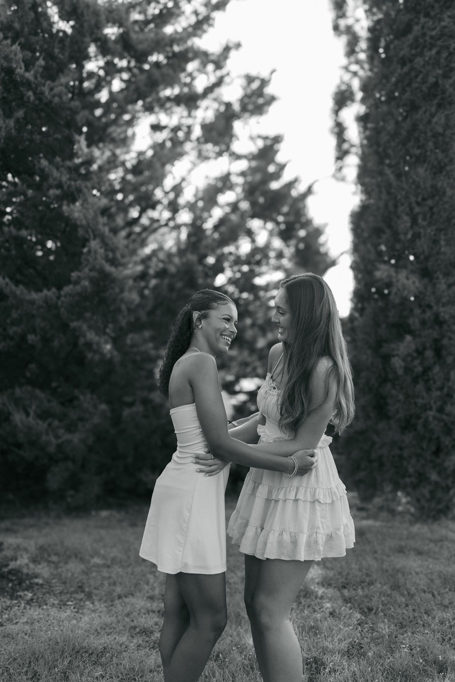 Two young women in white dresses laughing and embracing in a lush green park in black and white photo.