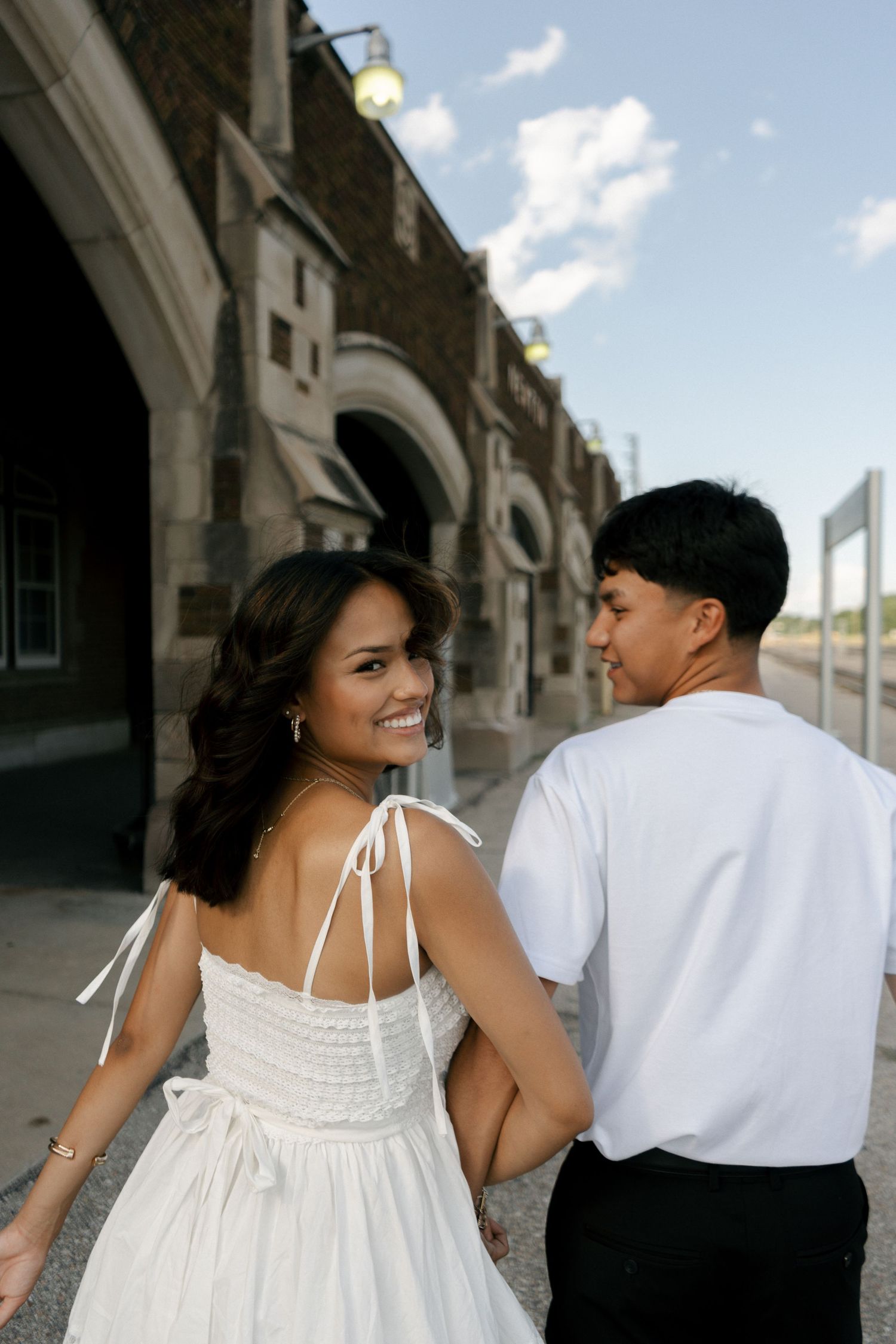 Smiling woman in white sundress walks hand-in-hand with man in white shirt along historic brick building.