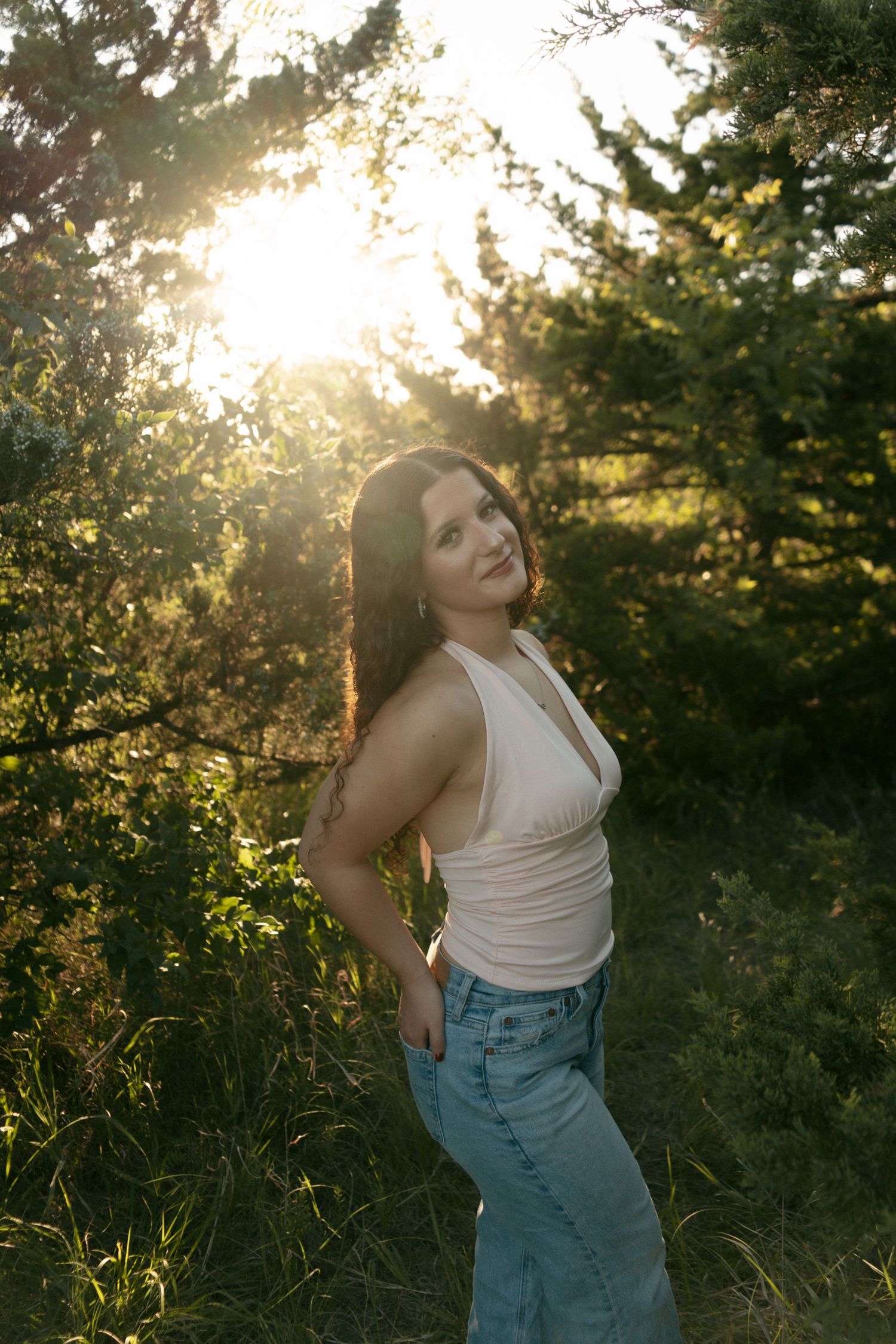 Woman in white tank top and jeans posing outdoors with golden sunset light through green trees behind her.