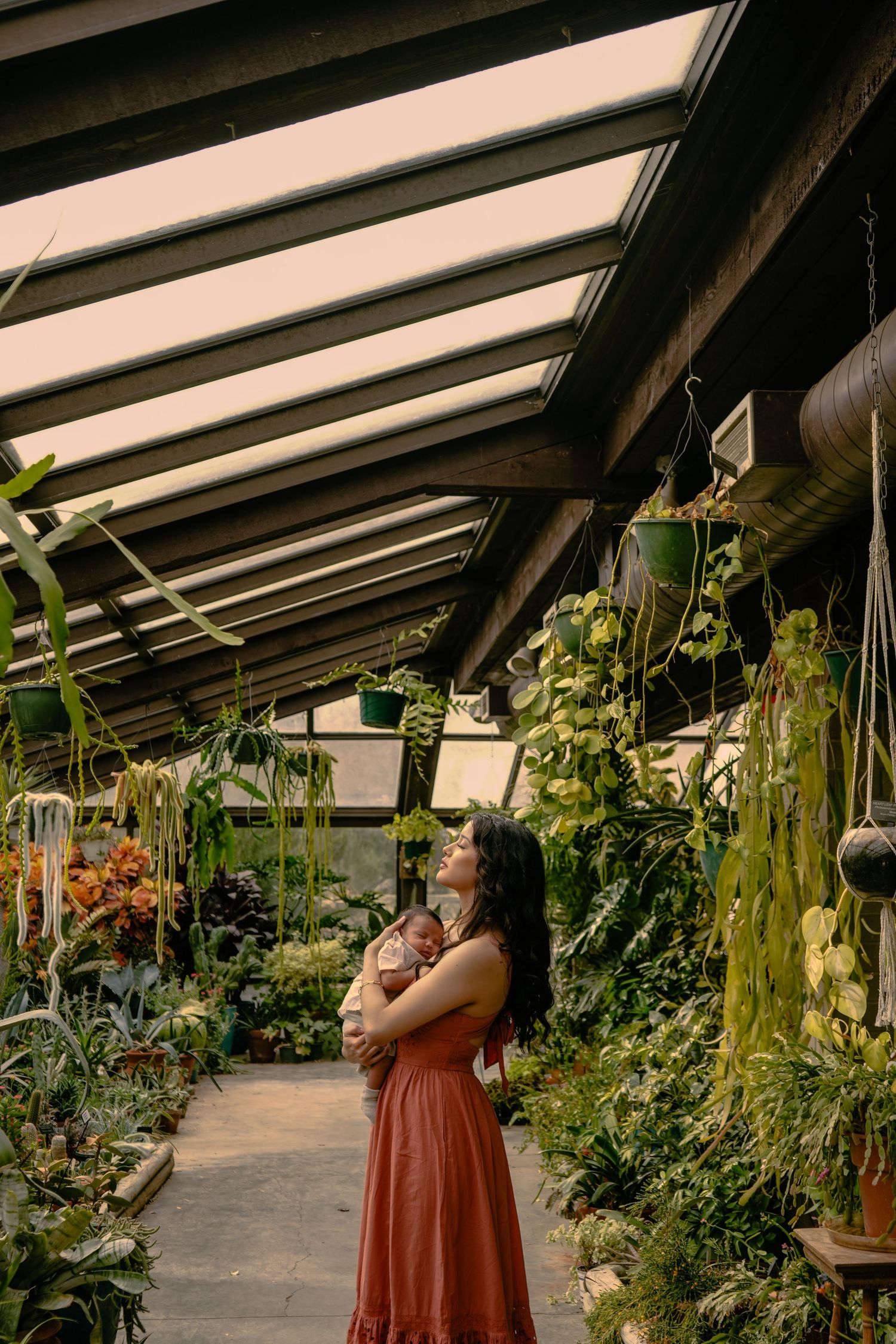 Woman in red dress cradling newborn baby in lush greenhouse with hanging plants and glass ceiling.