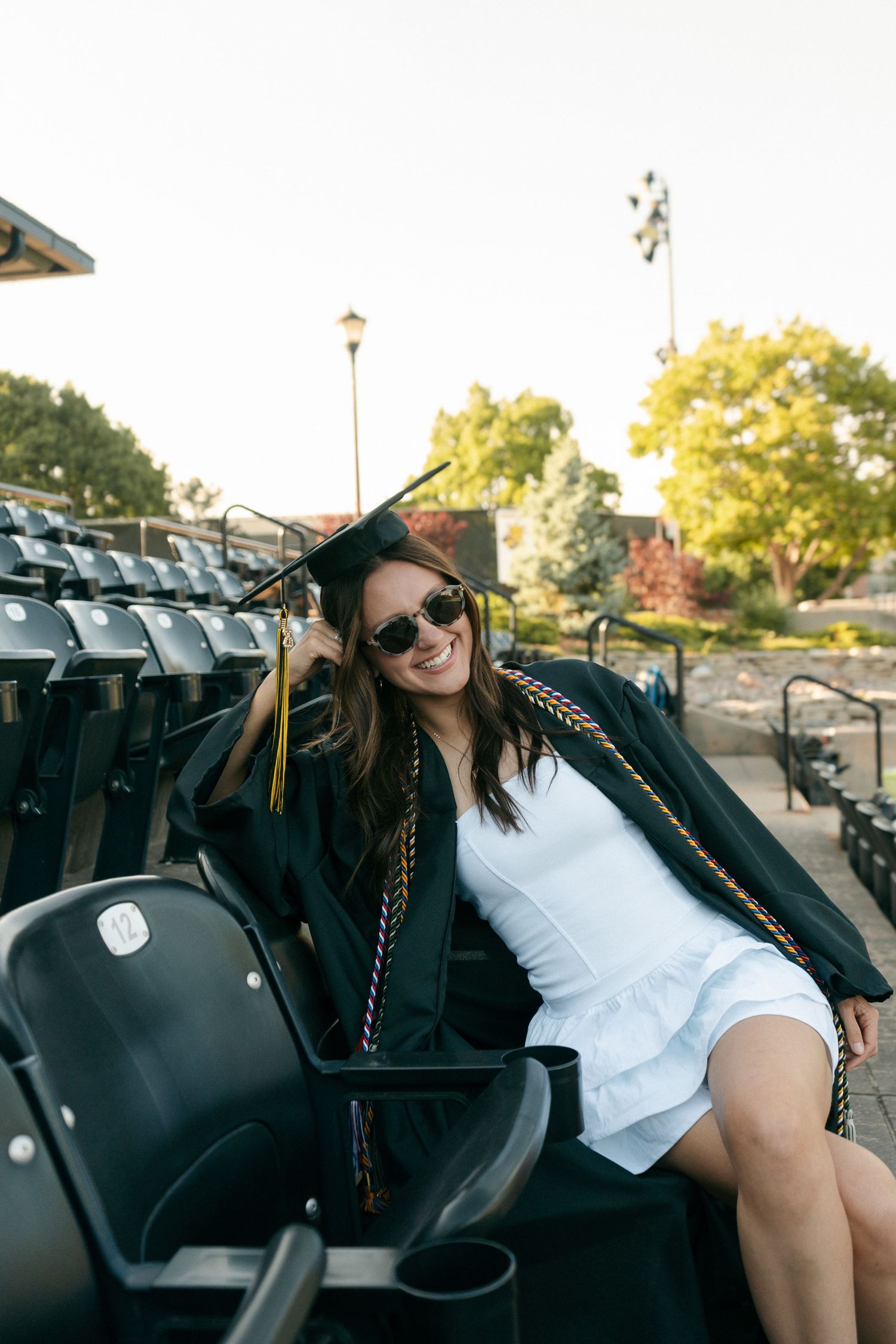 Smiling graduate in cap and gown with colorful tassels sits in stadium bleachers wearing white dress and sunglasses.
