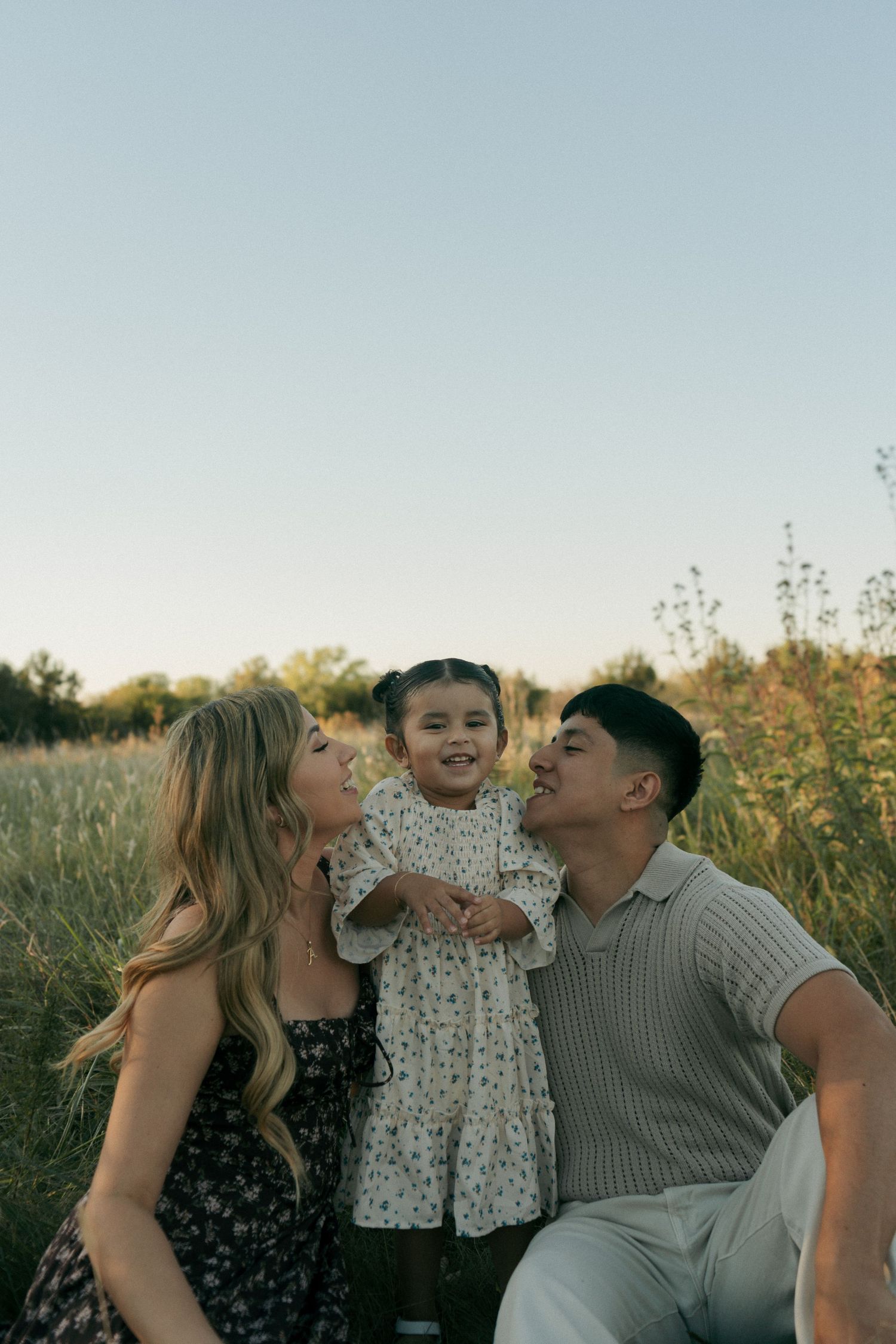 A smiling family of three sits in a sunlit meadow, with both parents looking lovingly at their happy toddler.