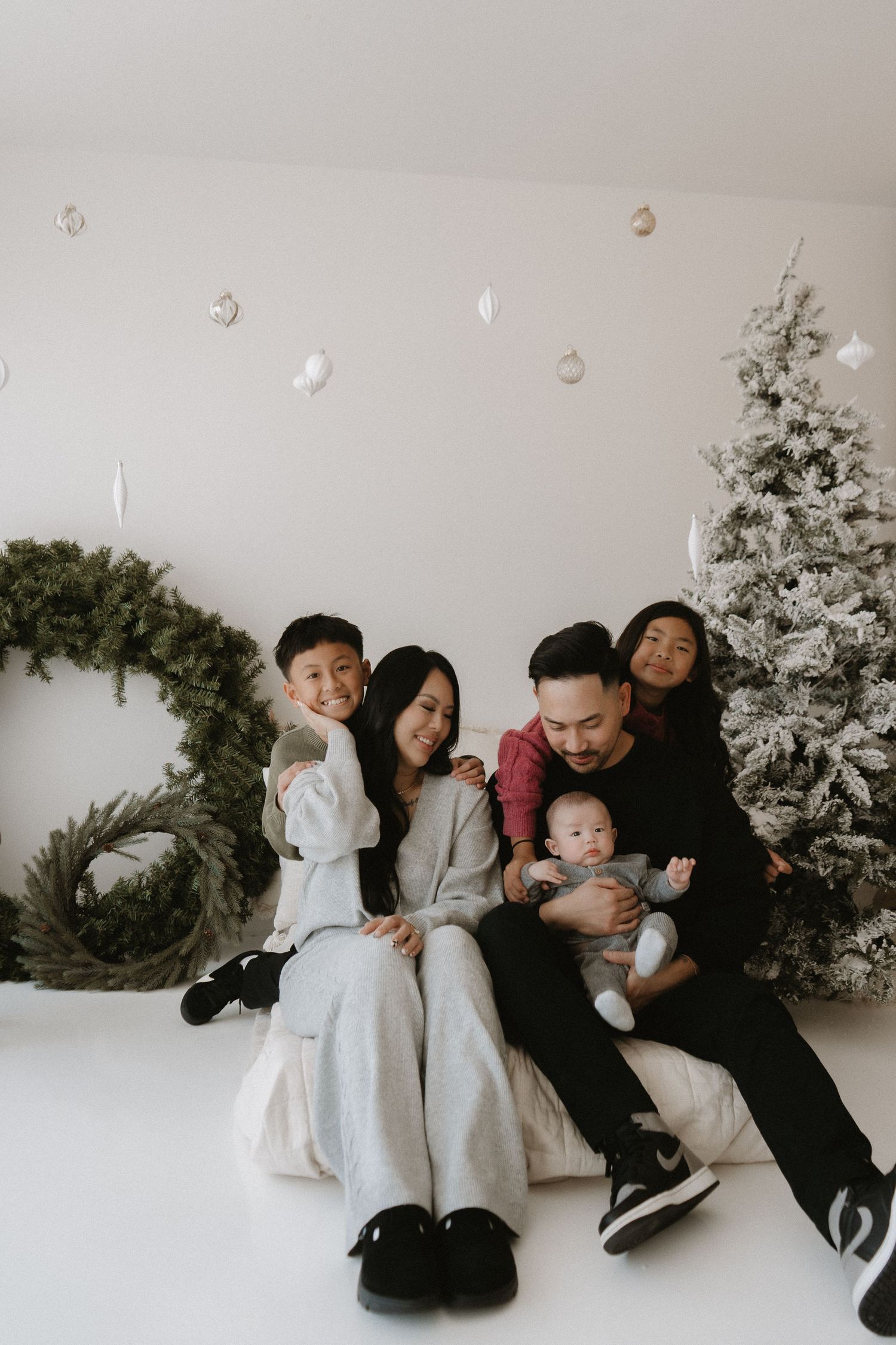 A joyful Asian family of five poses together for a Christmas portrait with a flocked tree and wreath.