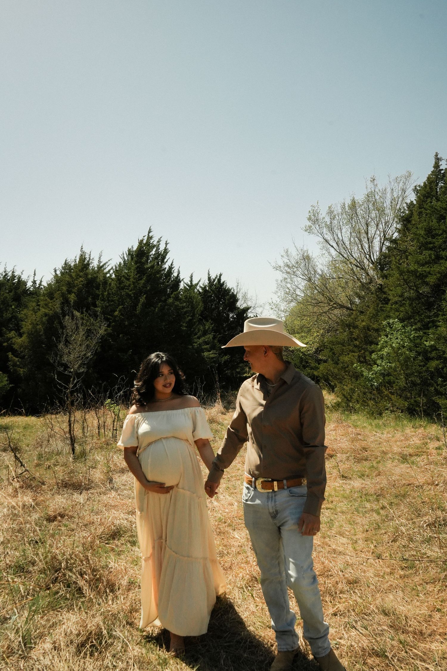Expectant couple holding hands in field, woman in cream maternity dress, man in cowboy hat and jeans.
