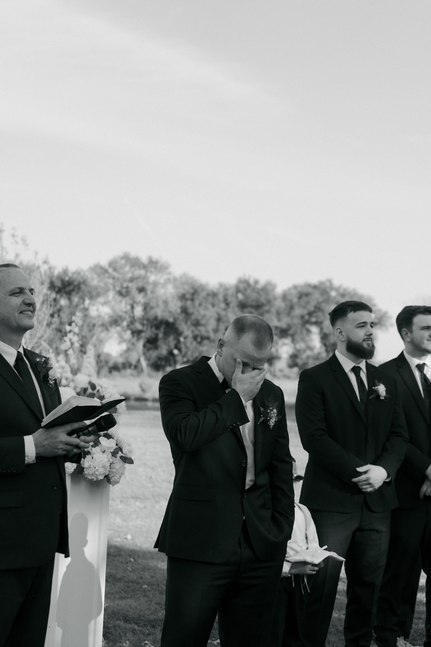 Emotional black and white photo of groom wiping tears at altar during outdoor wedding ceremony.