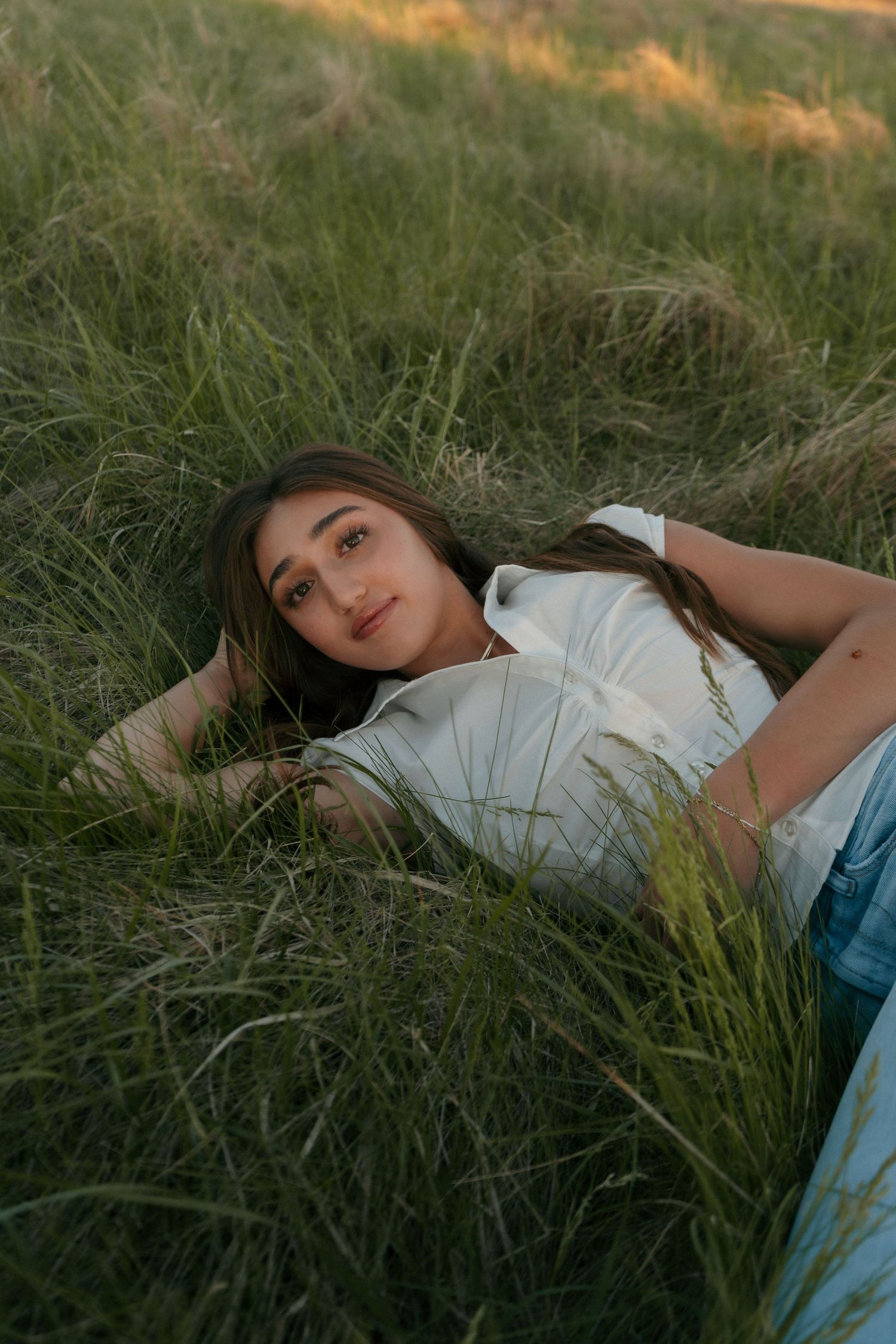 Young woman relaxing in lush green grass field, looking at camera during warm sunset lighting.