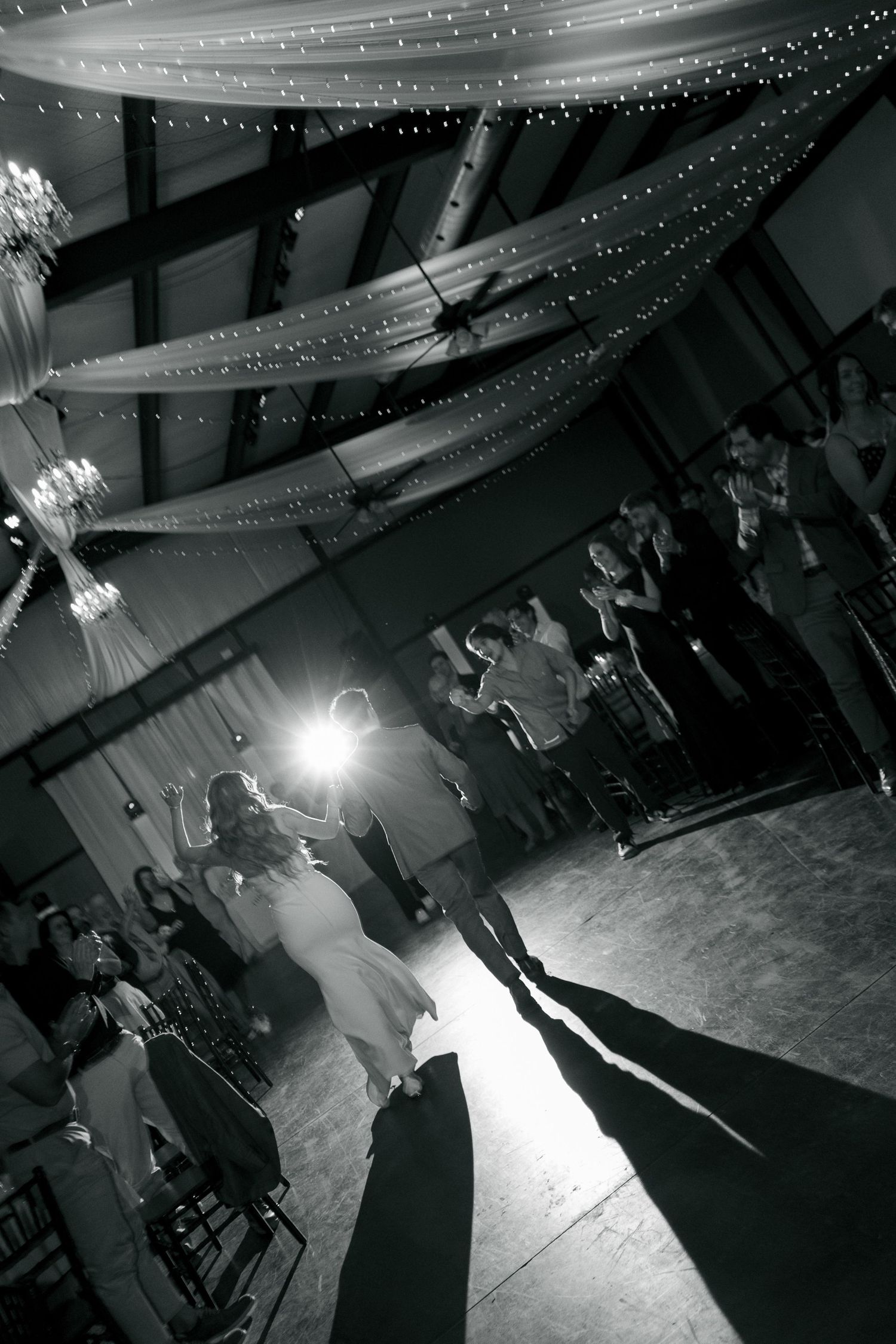 Black and white photo of couple sharing first dance at wedding reception with draped ceiling lights and guests watching.