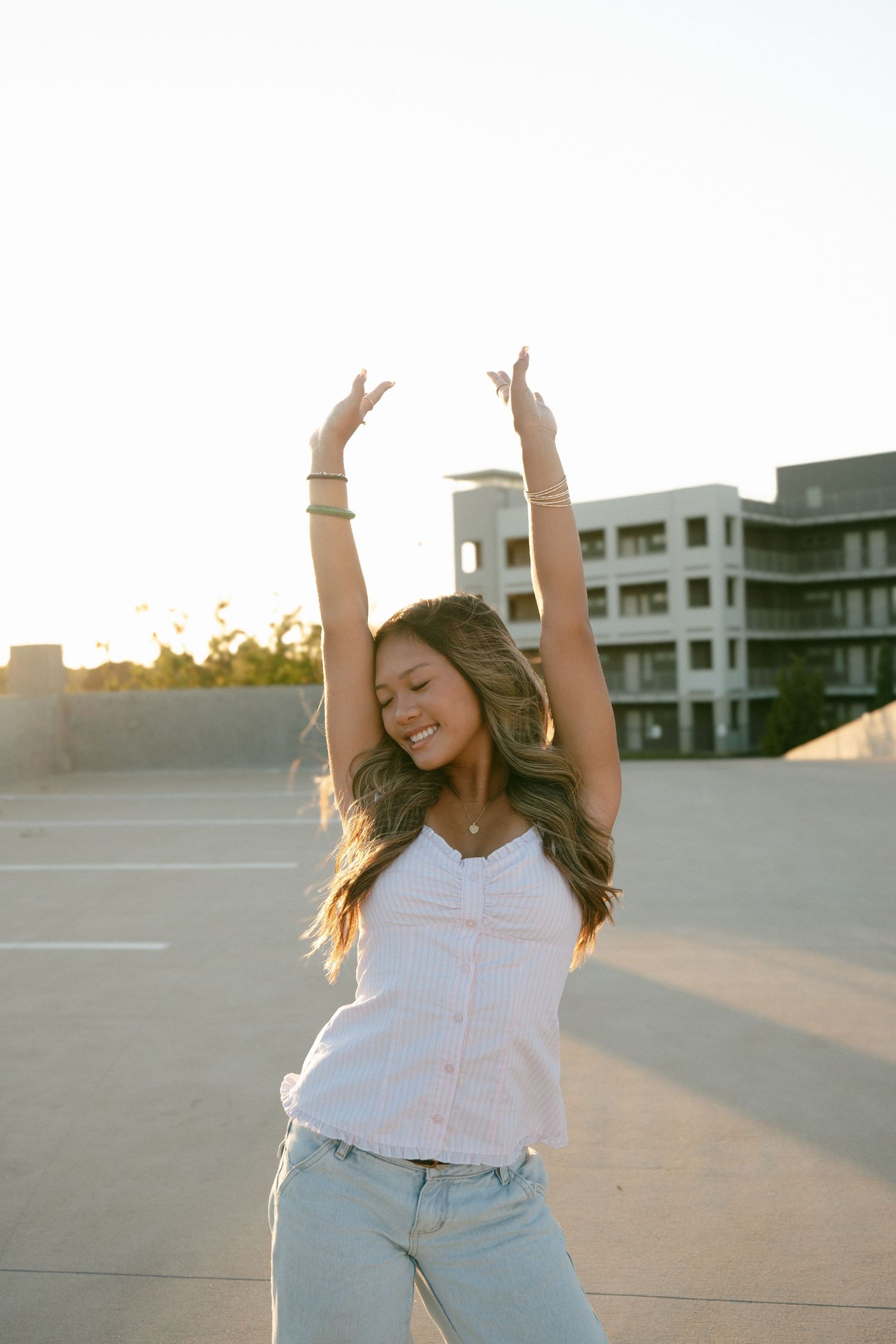 Joyful young woman in white top raising arms in empty parking lot during golden hour sunset.