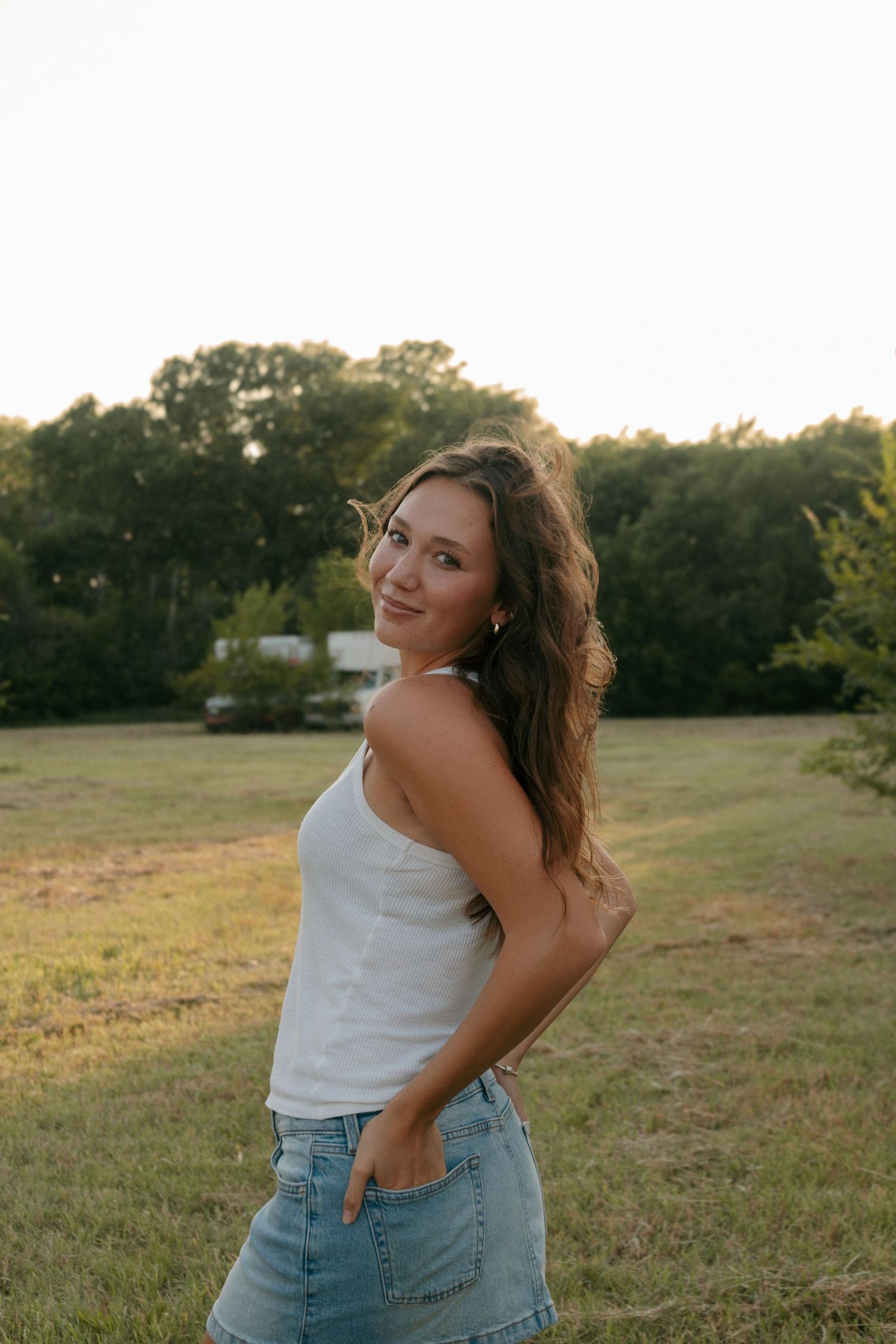 Young woman in white tank top and denim skirt smiling over shoulder in a sunlit grassy field.