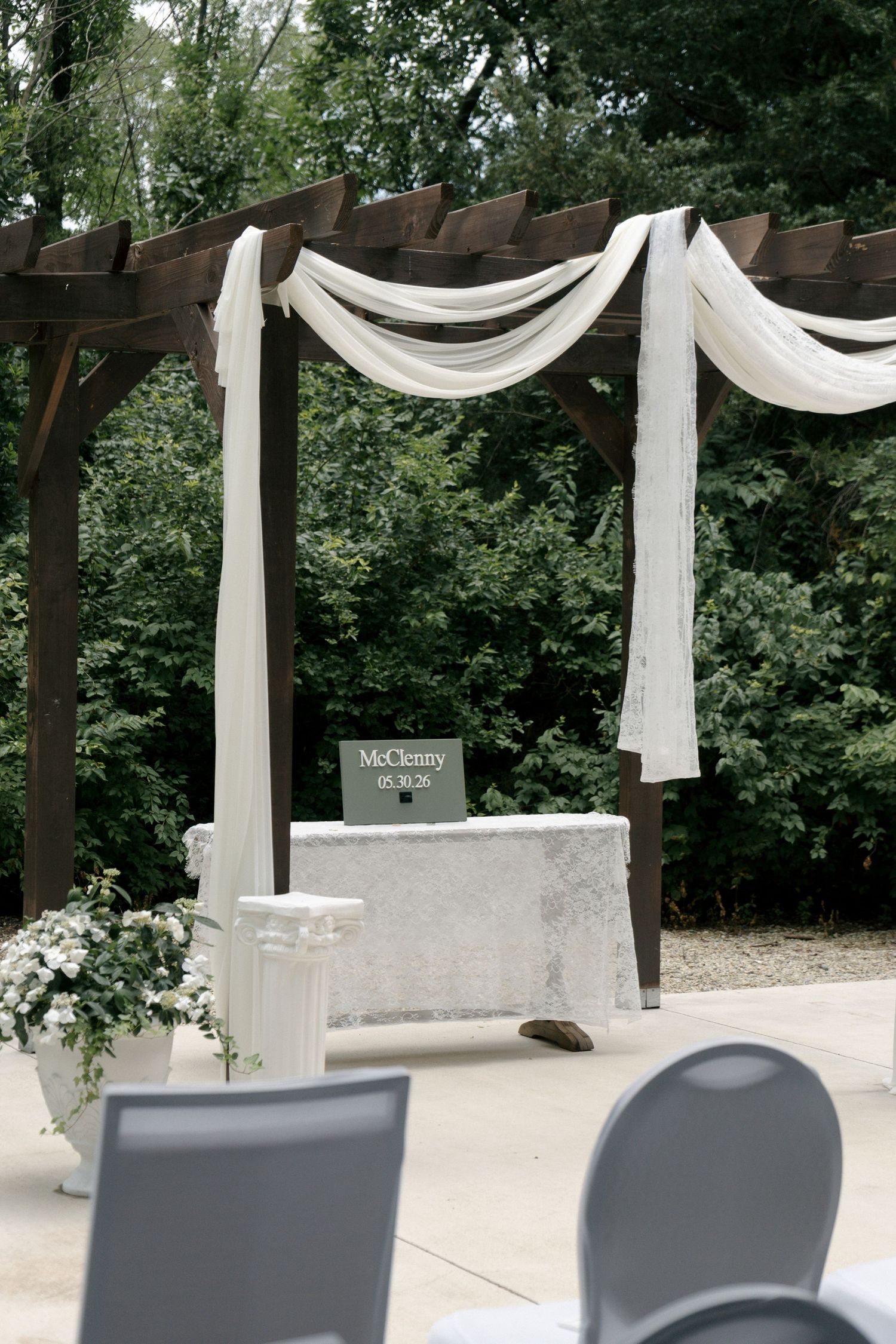 Outdoor wedding ceremony pergola draped with white fabric, featuring a decorated altar table with floral arrangements and signage.