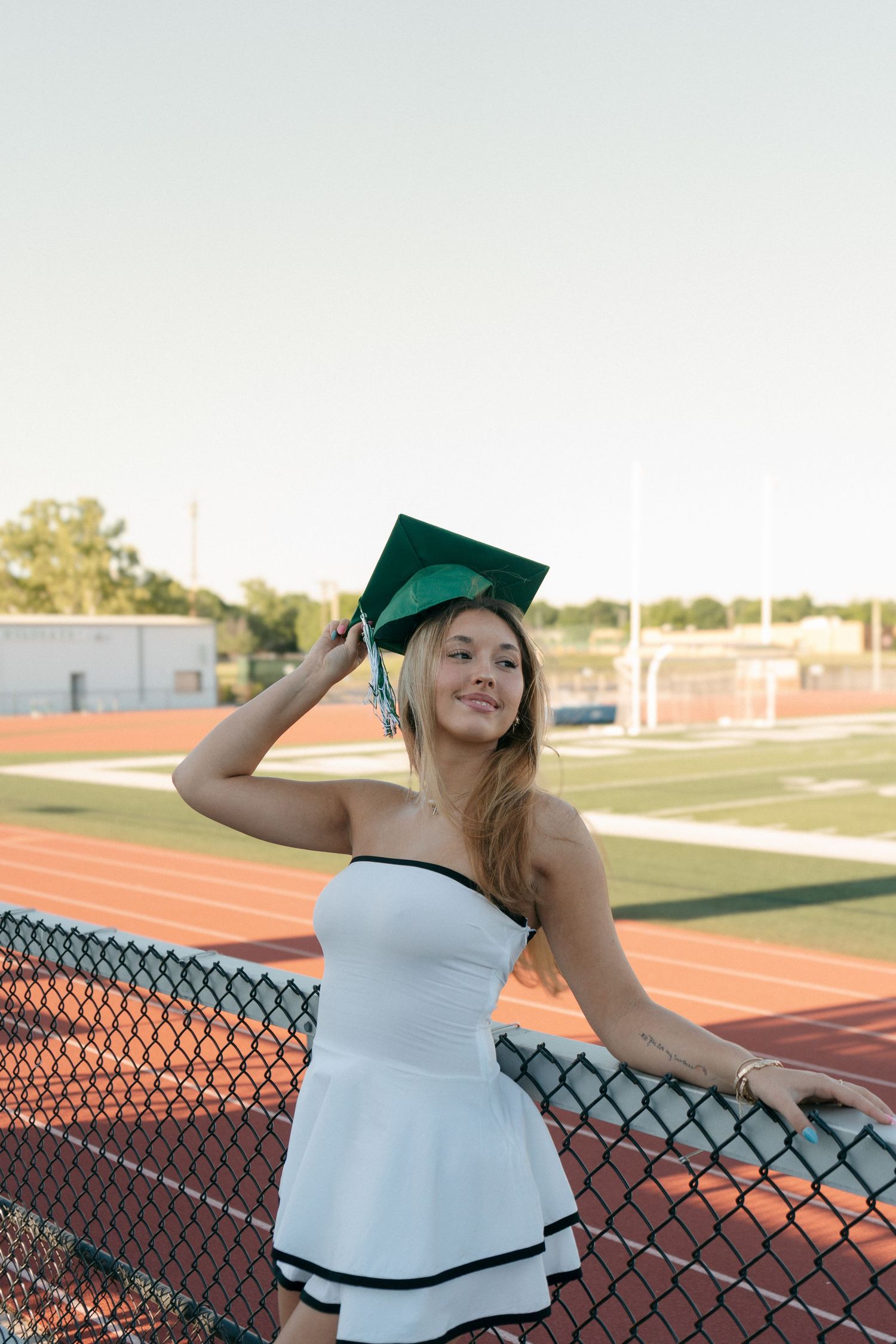 Smiling senior girl in white dress leans on fence wearing green grad cap.
