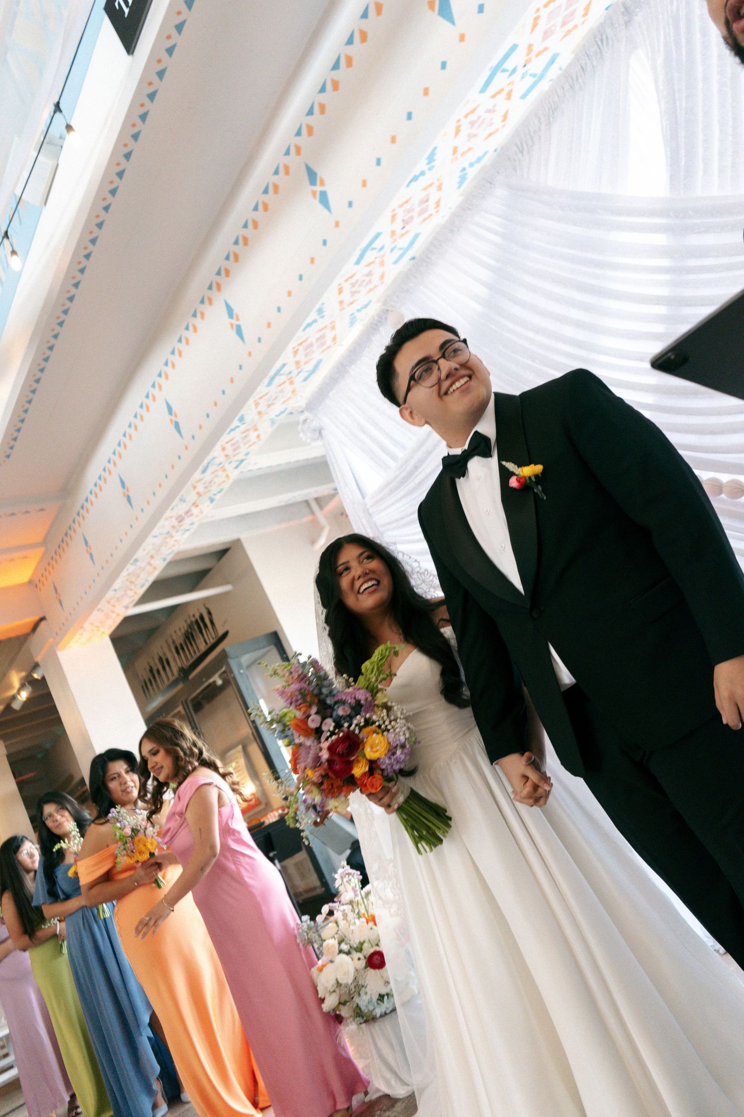 Joyful bride and groom walk down the aisle at wedding ceremony with colorful bridesmaids and floral bouquet.