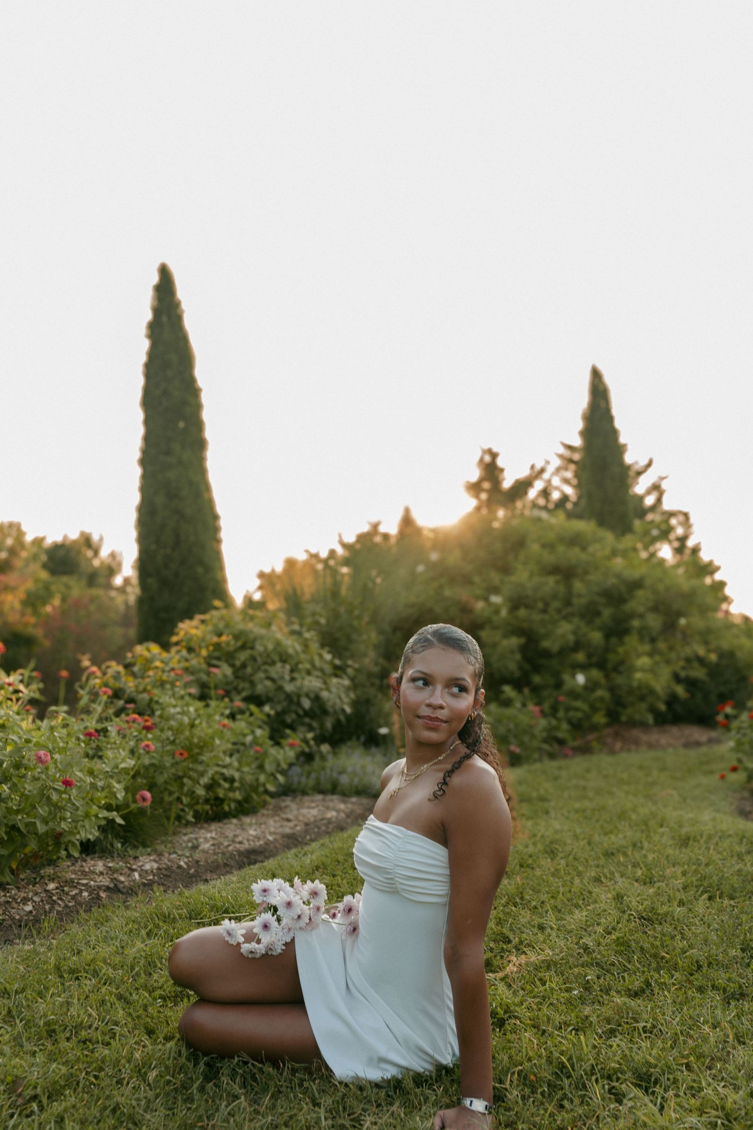 Young woman in white mini dress sitting on grass holding flowers in a garden at sunset.