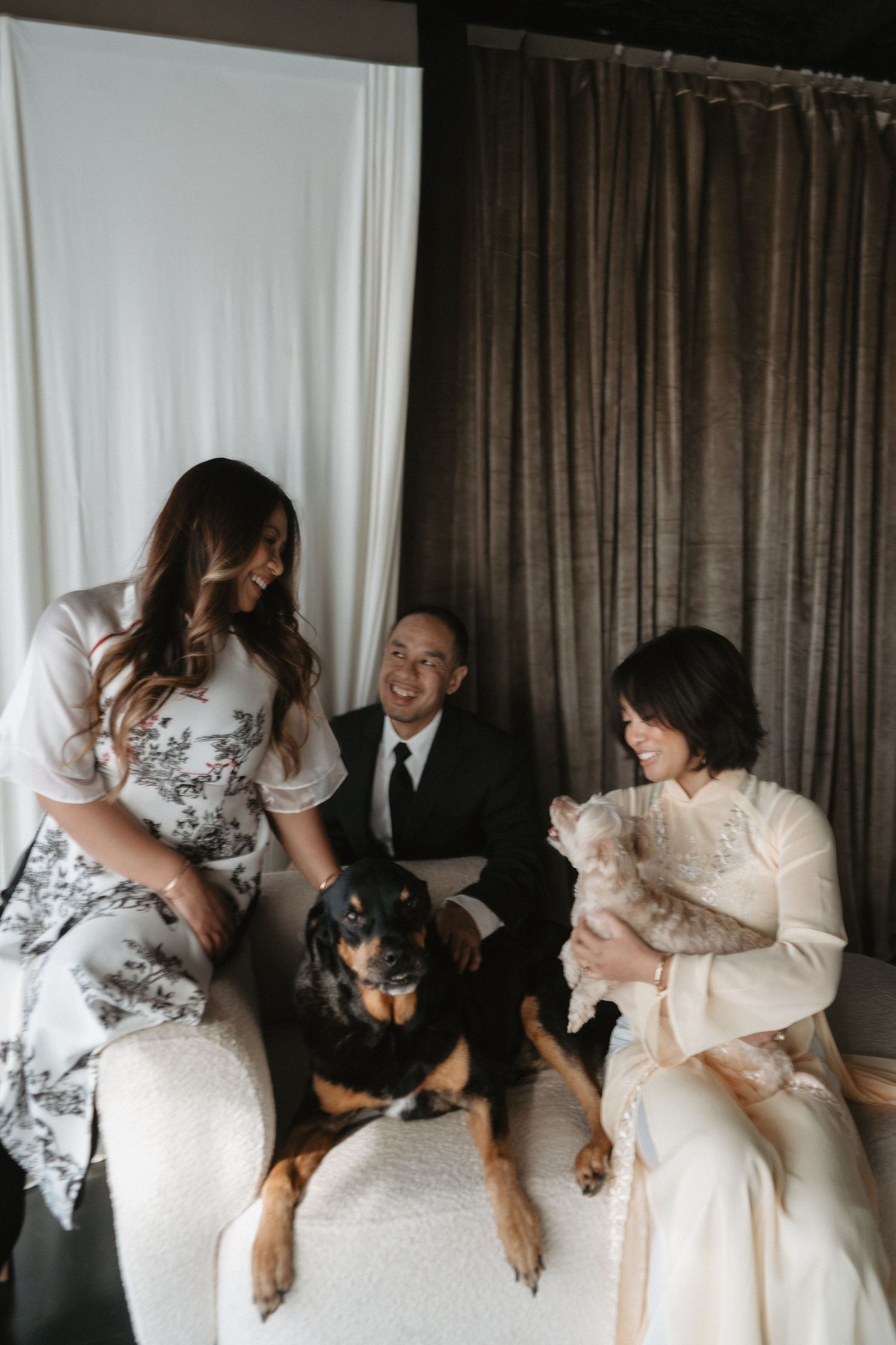 A bride in a white gown and groom in a suit pose with a woman and their Rottweiler dog.