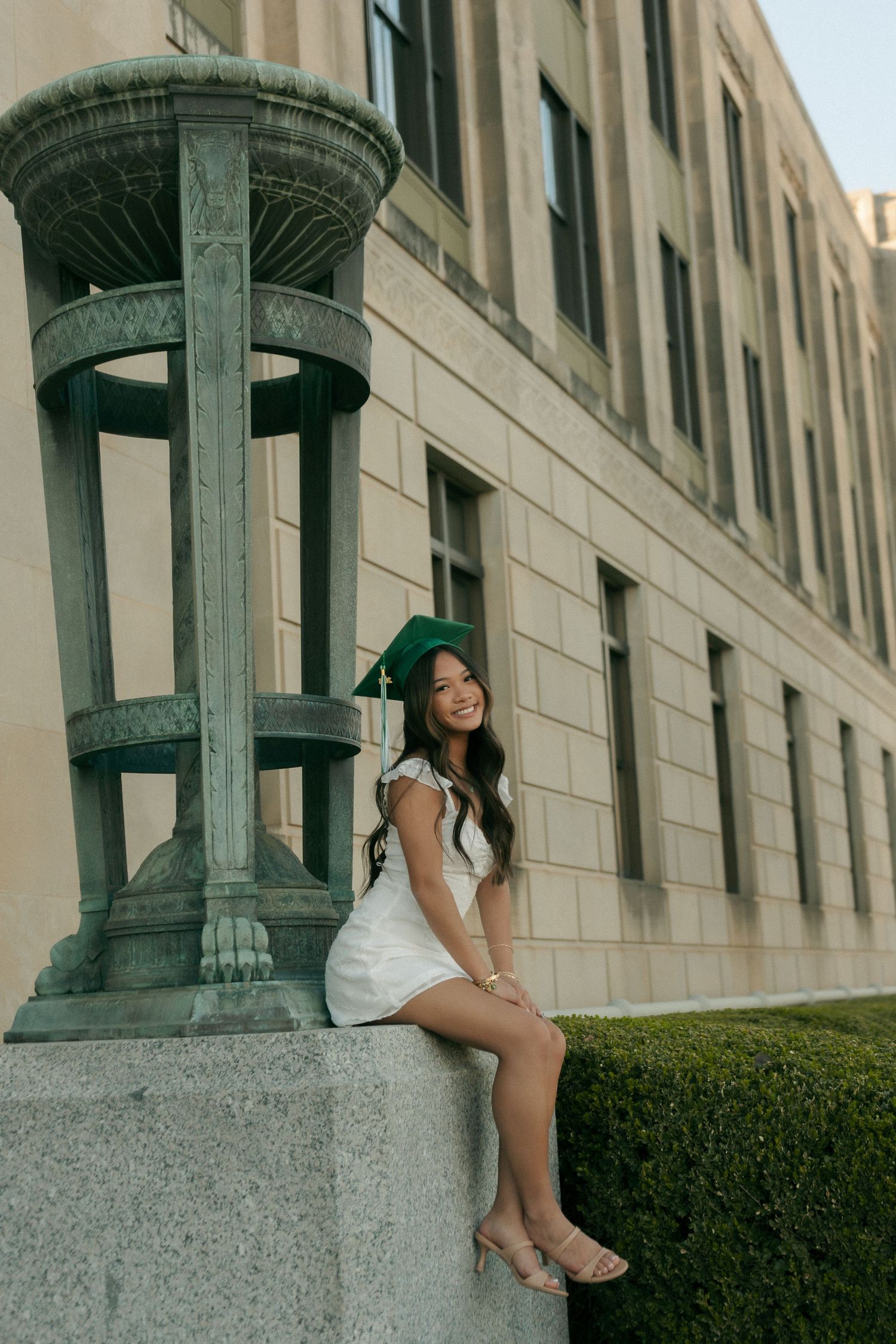 Smiling graduate in green cap and white dress sitting on stone wall beside decorative bronze column.