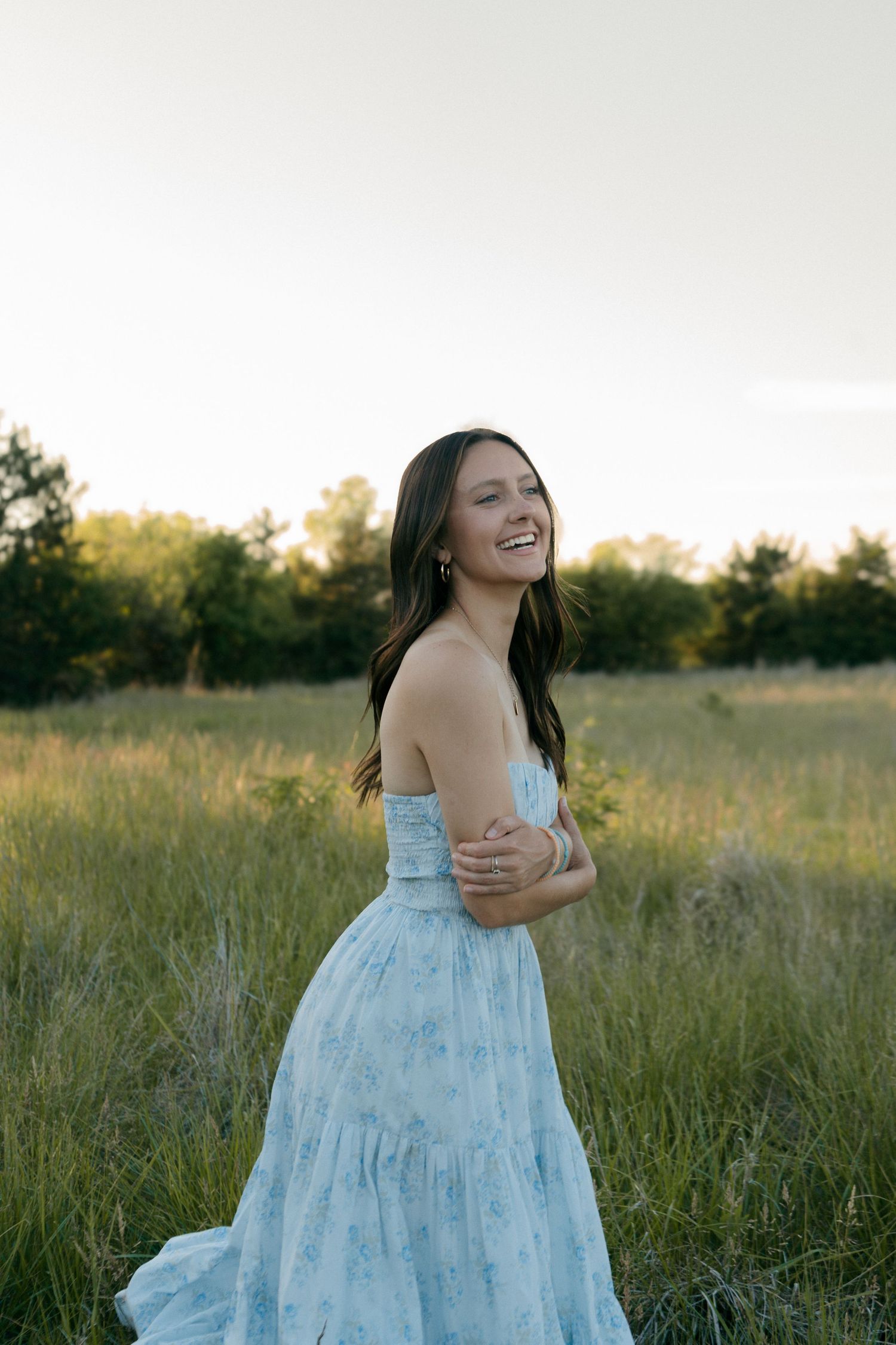 Smiling young woman in a light blue floral strapless dress standing in a green meadow at golden hour.