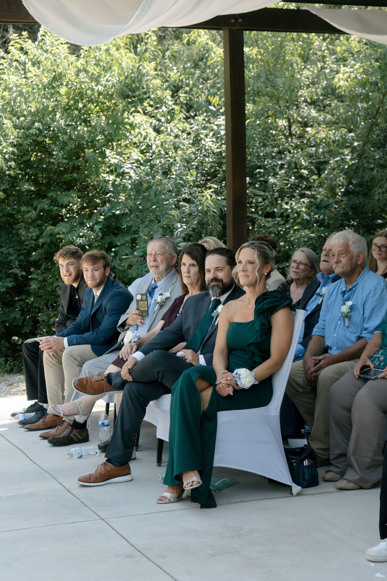 Guests seated outdoors at a wedding ceremony, wearing formal attire, surrounded by lush green trees.