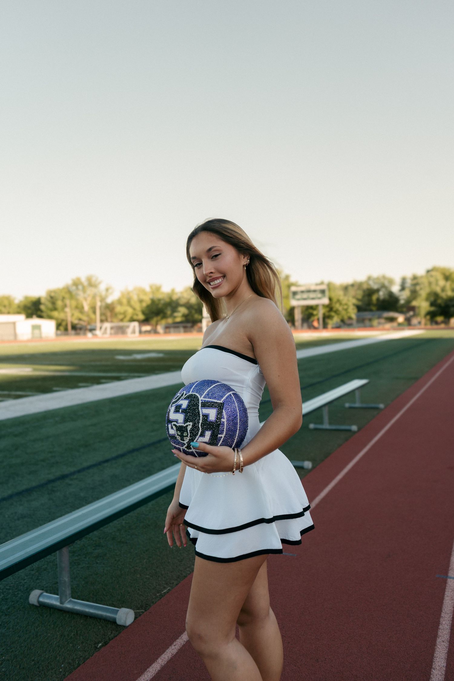 Smiling senior girl in white dress holds school volleyball on running track.