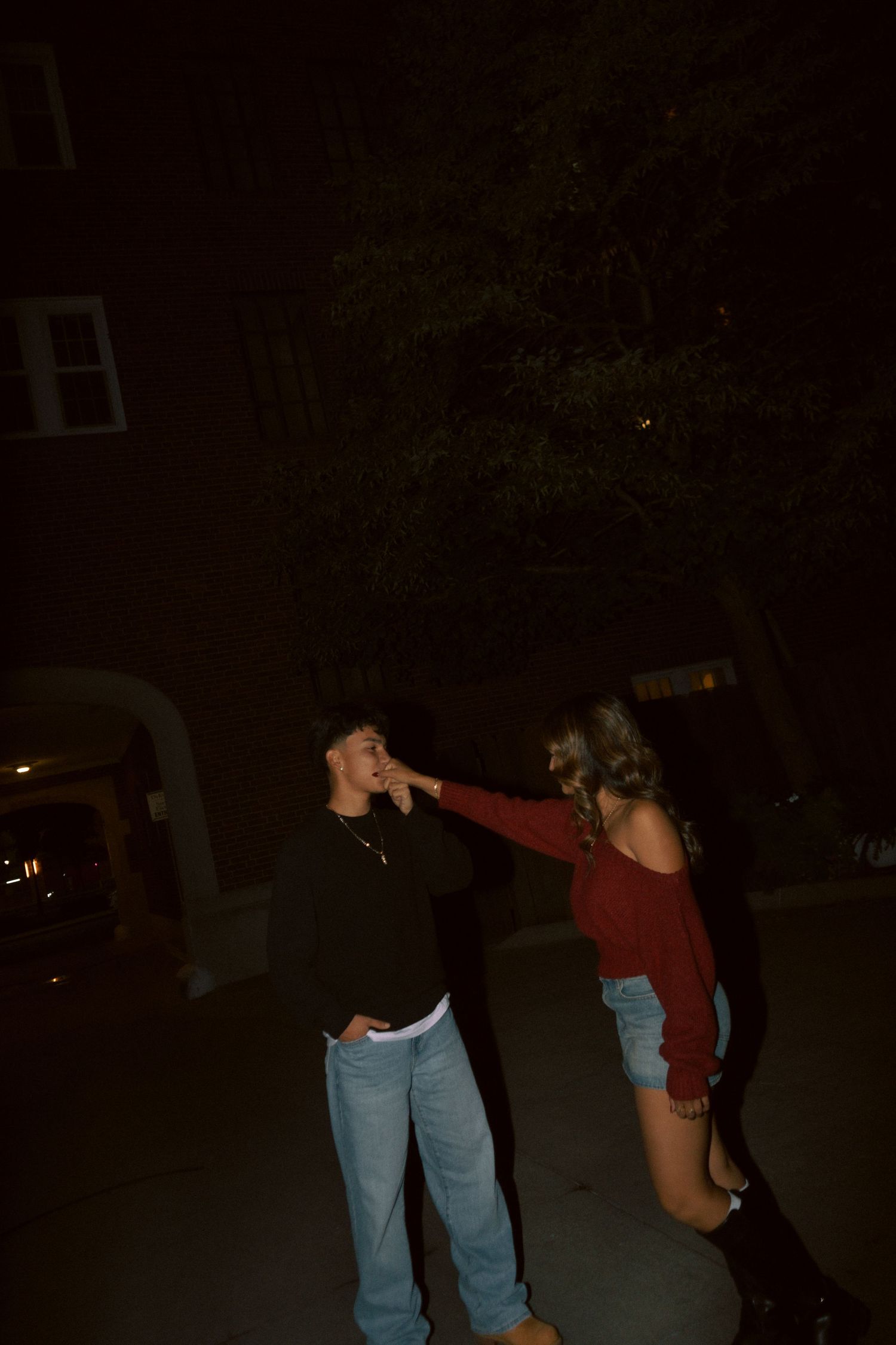 A young couple shares a playful moment outdoors at night, with the woman touching the man's face.