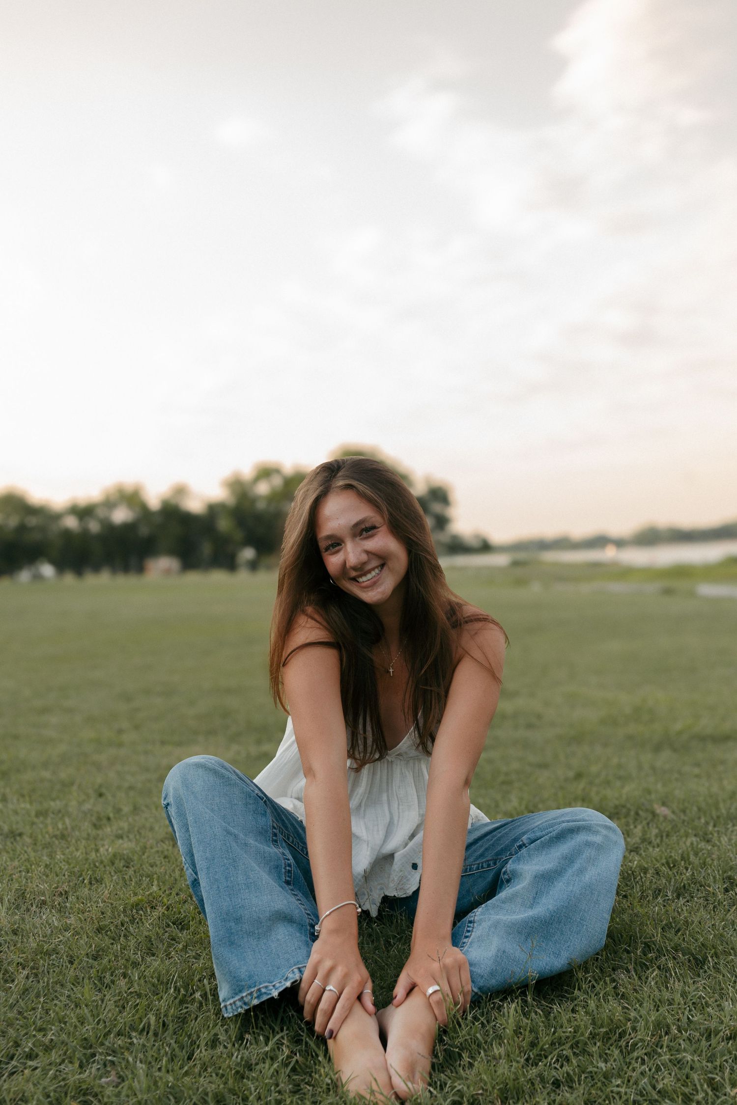 Laughing young woman in white camisole and jeans sits barefoot on a lush green lawn at dusk.