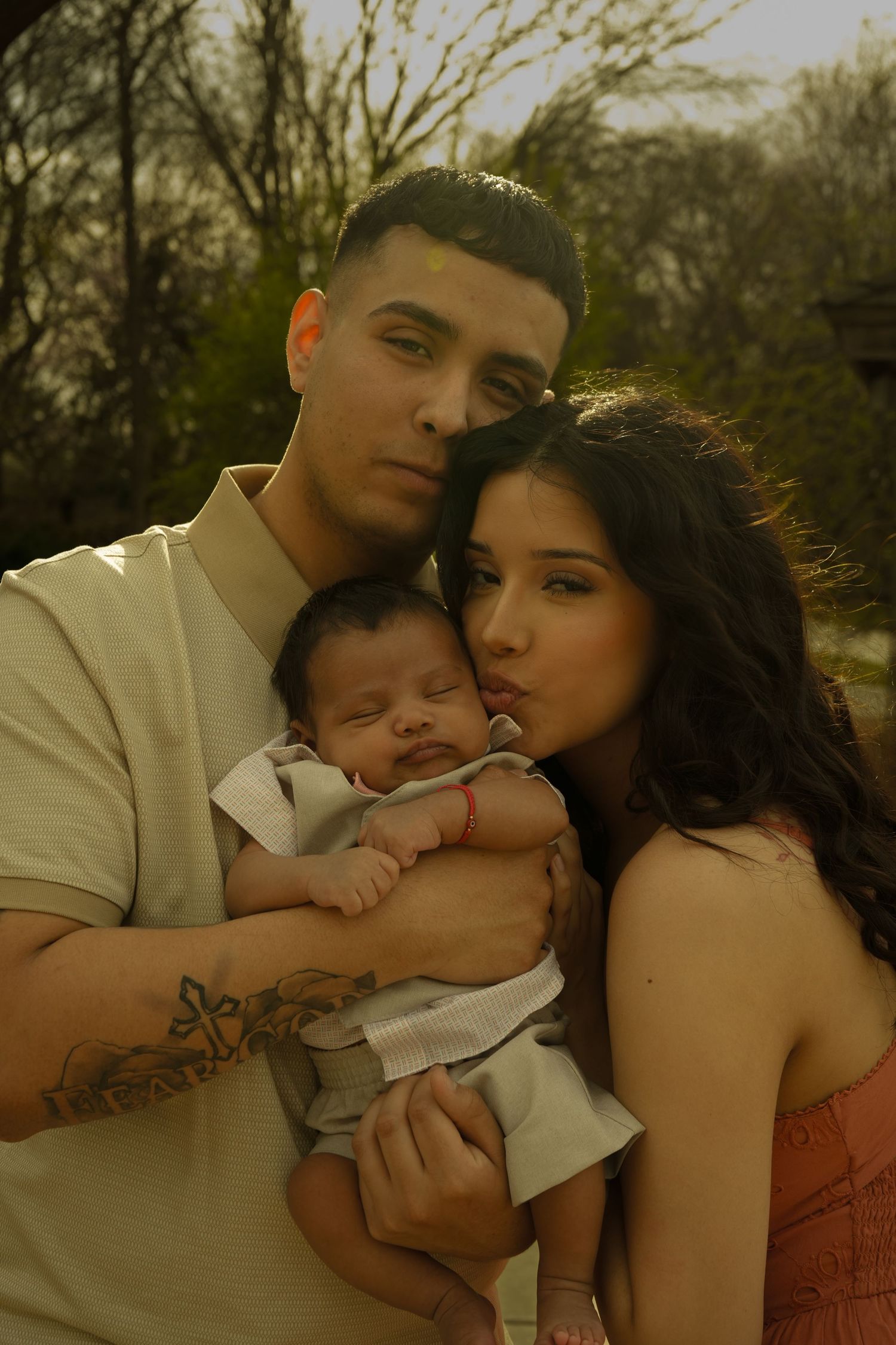 Young parents tenderly kissing newborn baby they hold together in outdoor golden hour portrait.