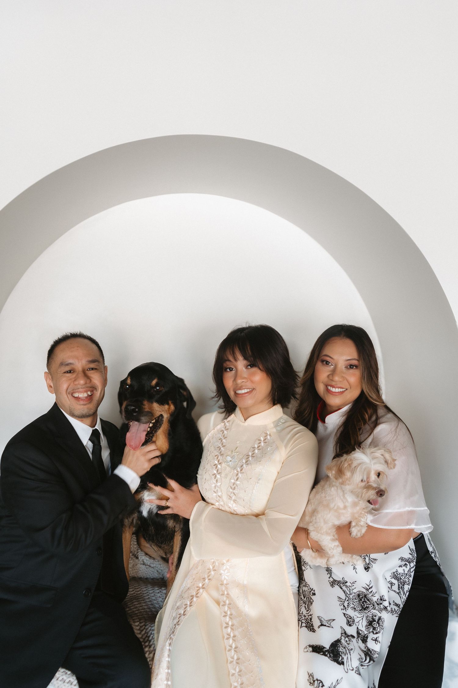 A bride in a traditional ao dai and groom pose with a woman and their two dogs under an arch.