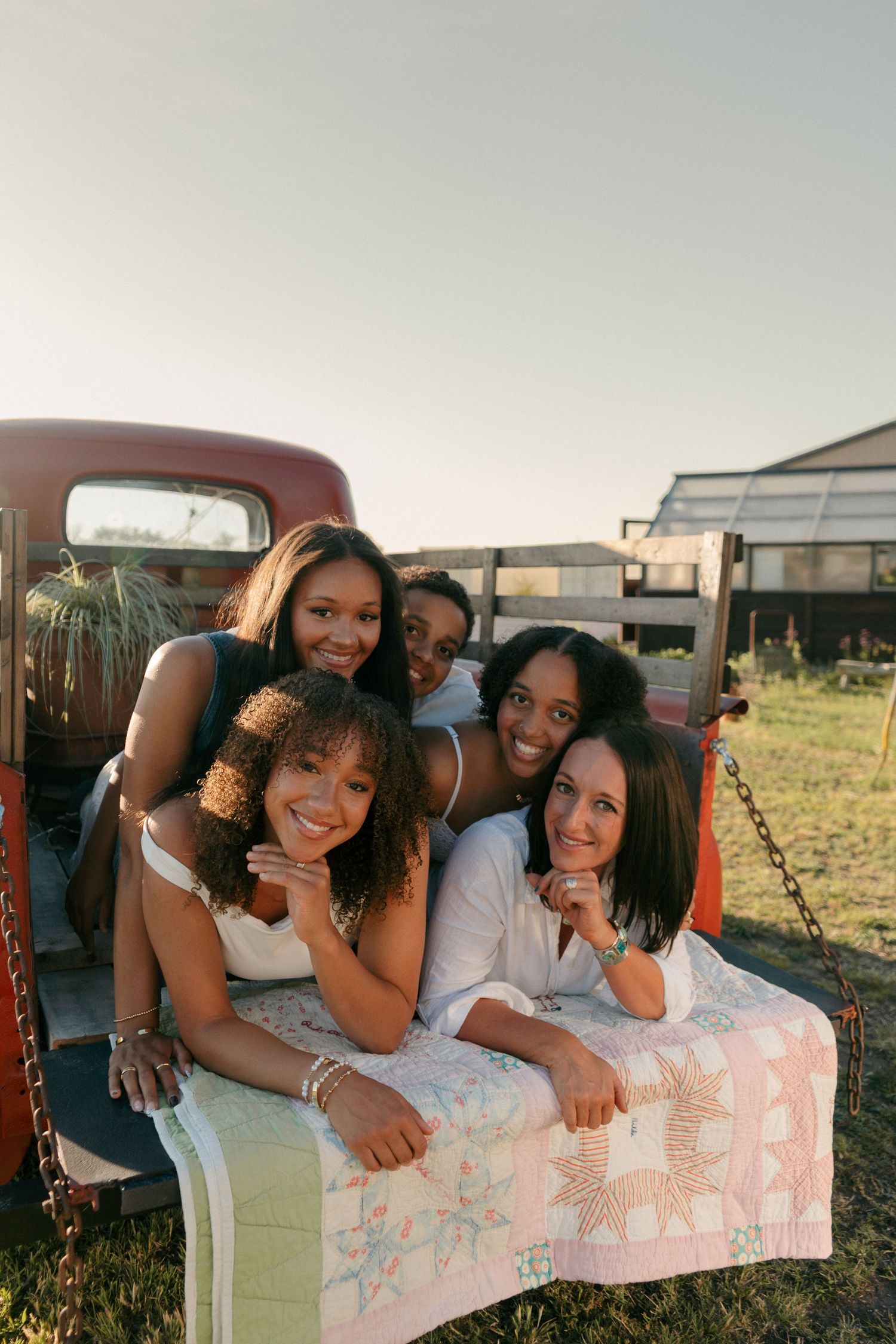 Five young women smiling together leaning on a vintage red truck tailgate with a quilt outdoors.