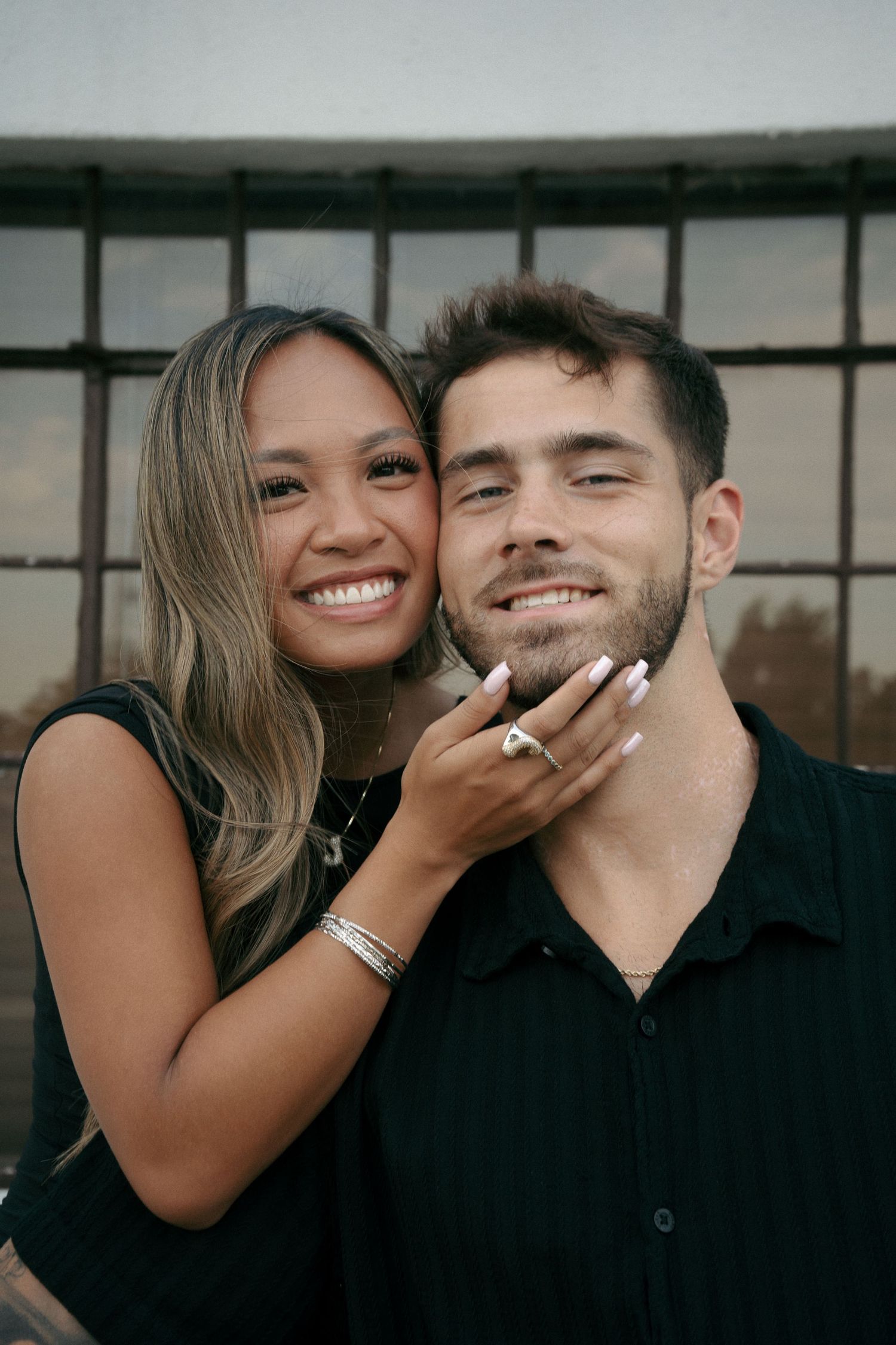 Smiling couple posing together, woman with long hair embracing man in black shirt near industrial window.
