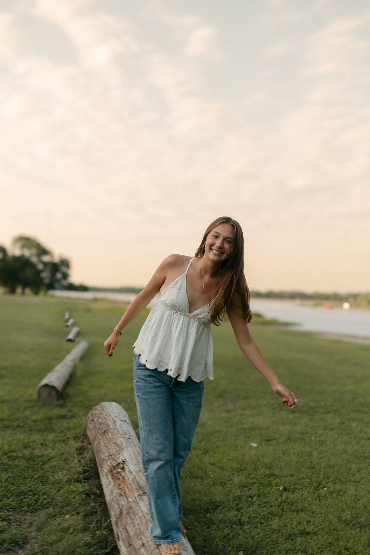 Laughing young woman in white camisole and jeans balancing on a log near a serene waterfront at dusk.