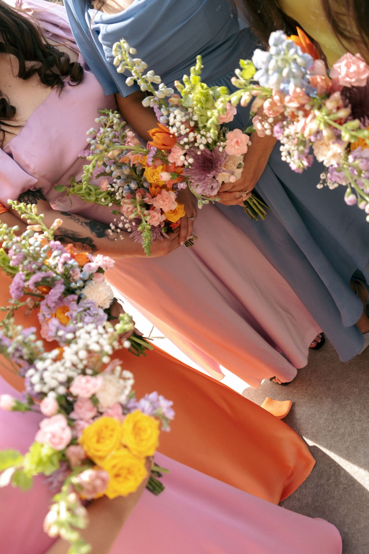 Bridesmaids in pink and blue gowns holding colorful wedding bouquets with roses, wildflowers, and mixed blooms outdoors.