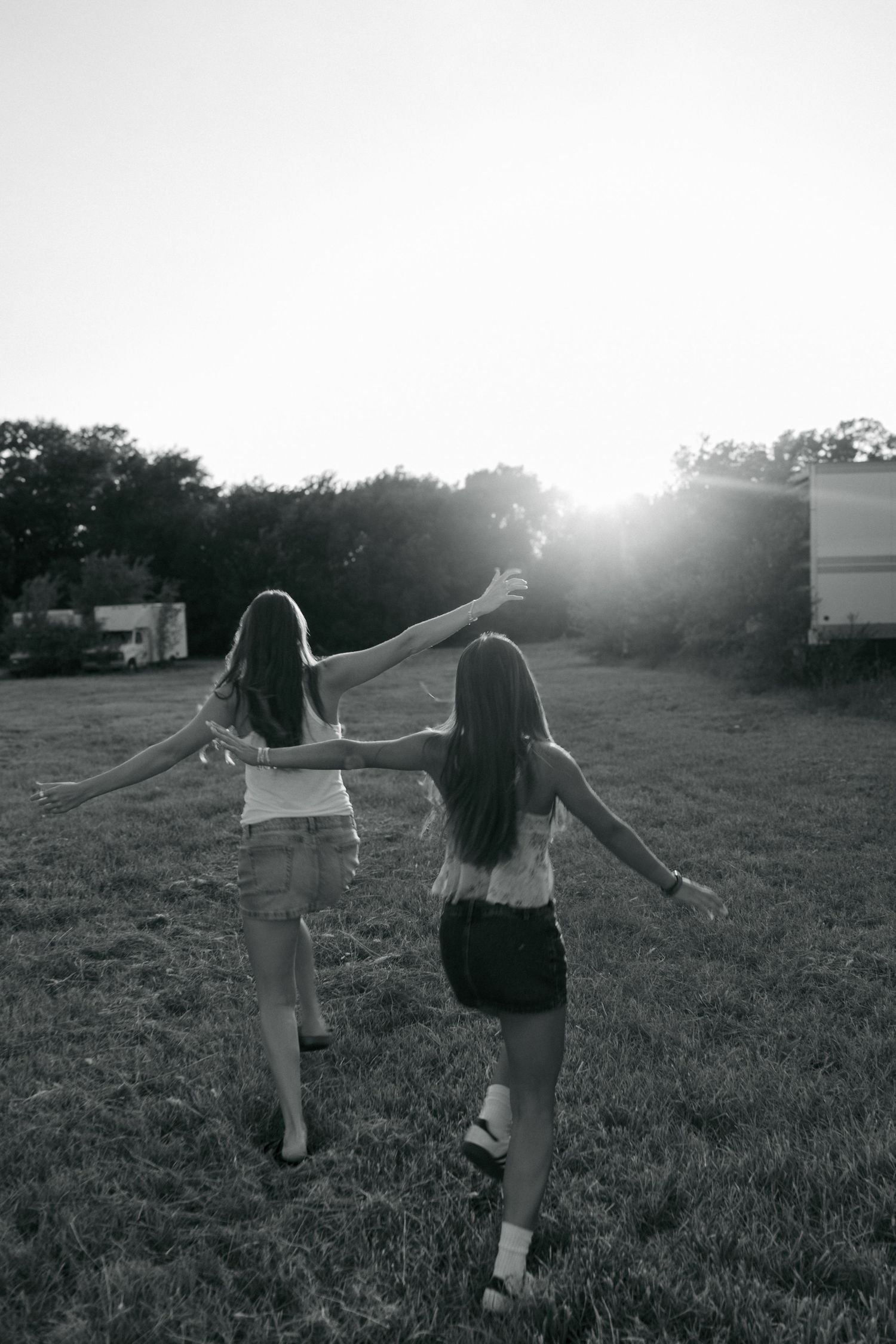 Black and white photo of two girls walking through a field with arms outstretched at sunset.