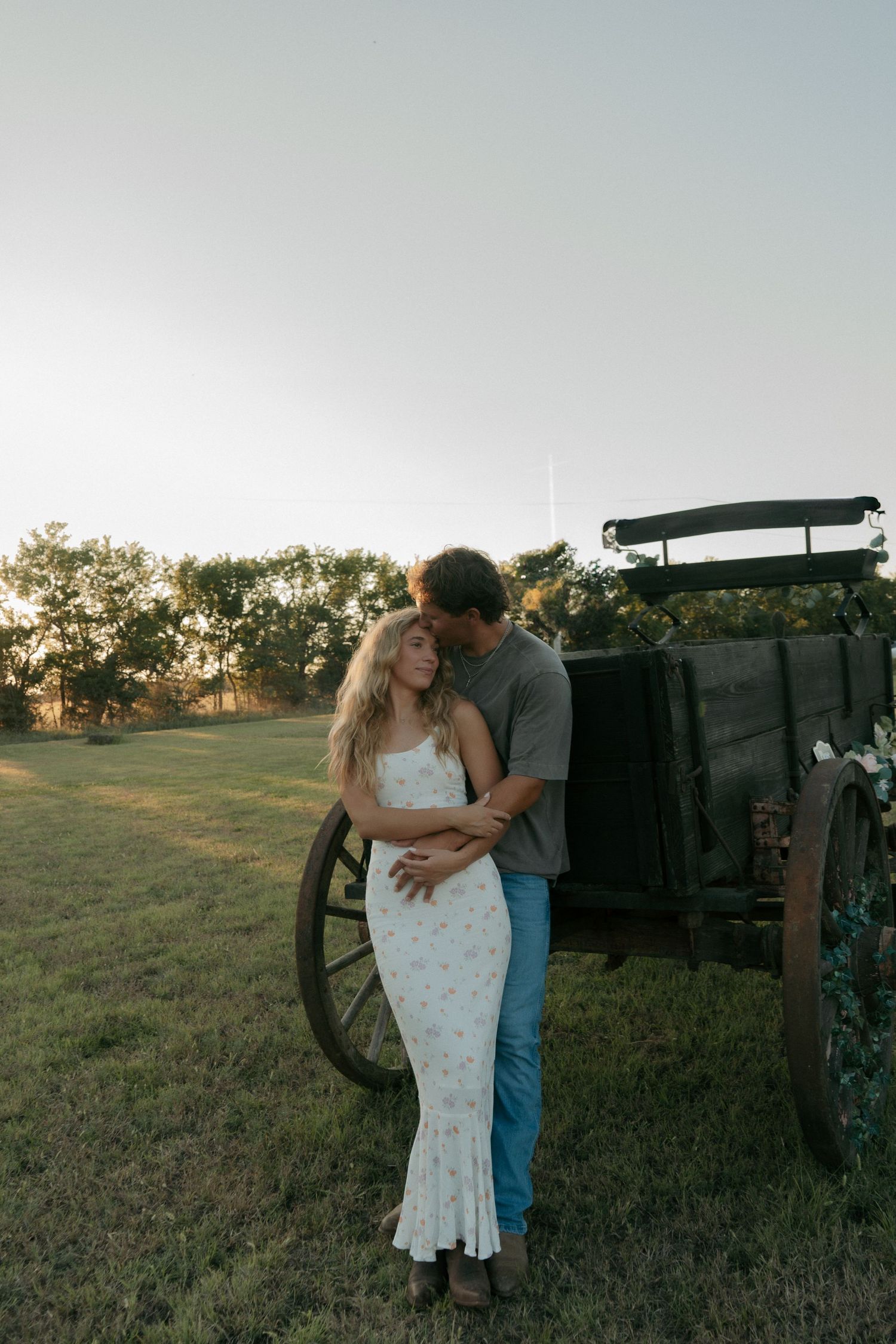 Couple sharing a tender embrace near a rustic wooden wagon decorated with flowers at sunset.
