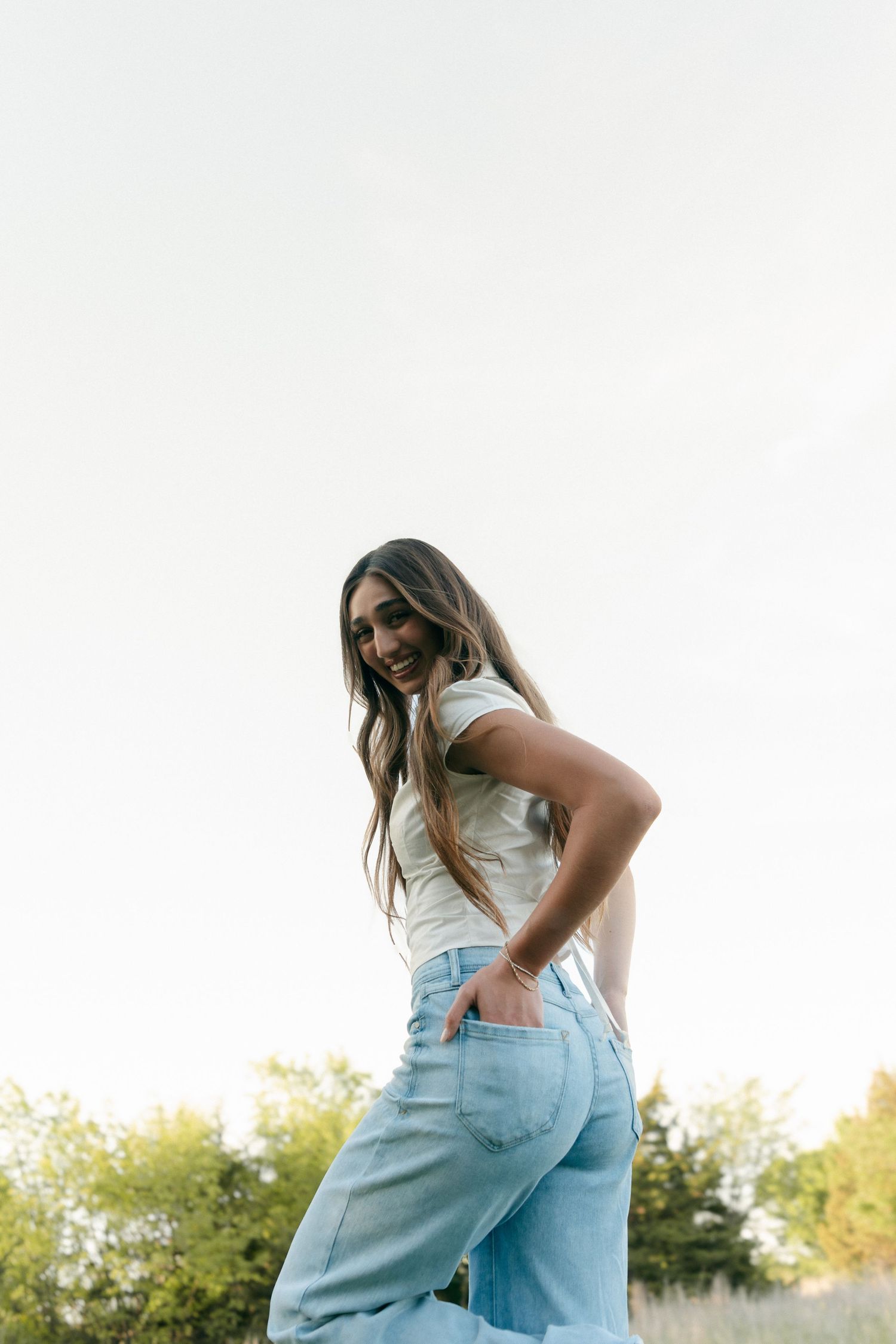 Smiling young woman in white top and light blue jeans posing outdoors against a bright sky.