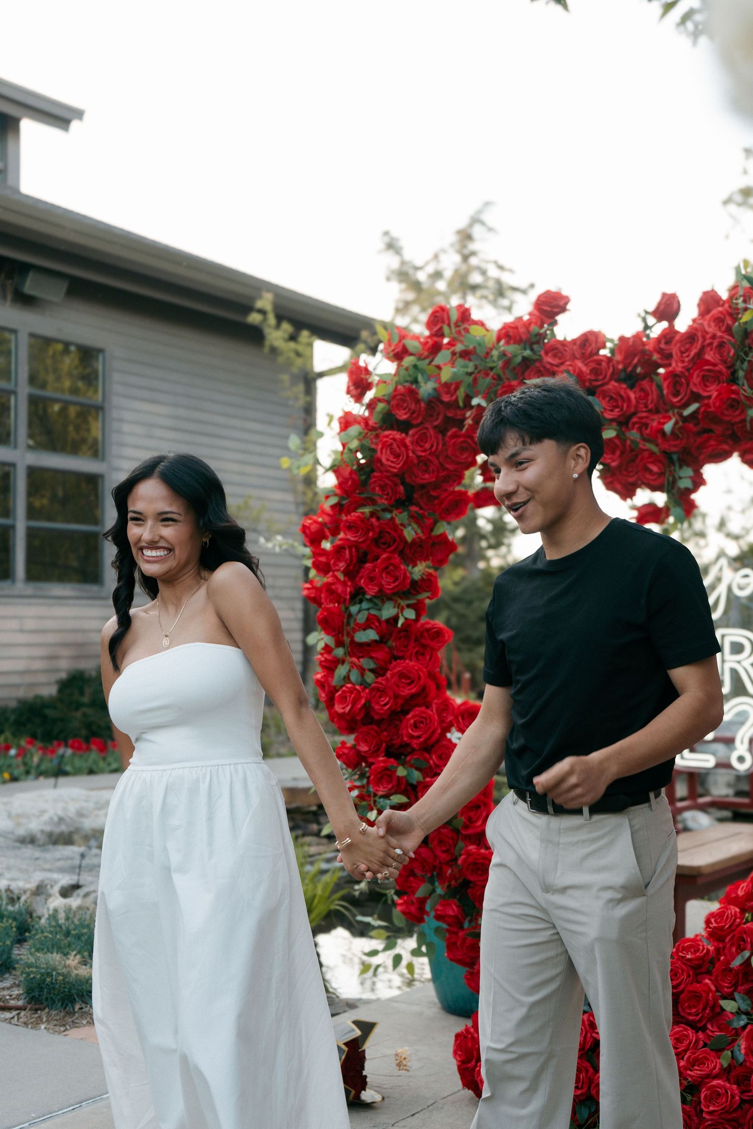 Joyful couple holding hands and laughing in front of red rose floral arch during proposal setup.