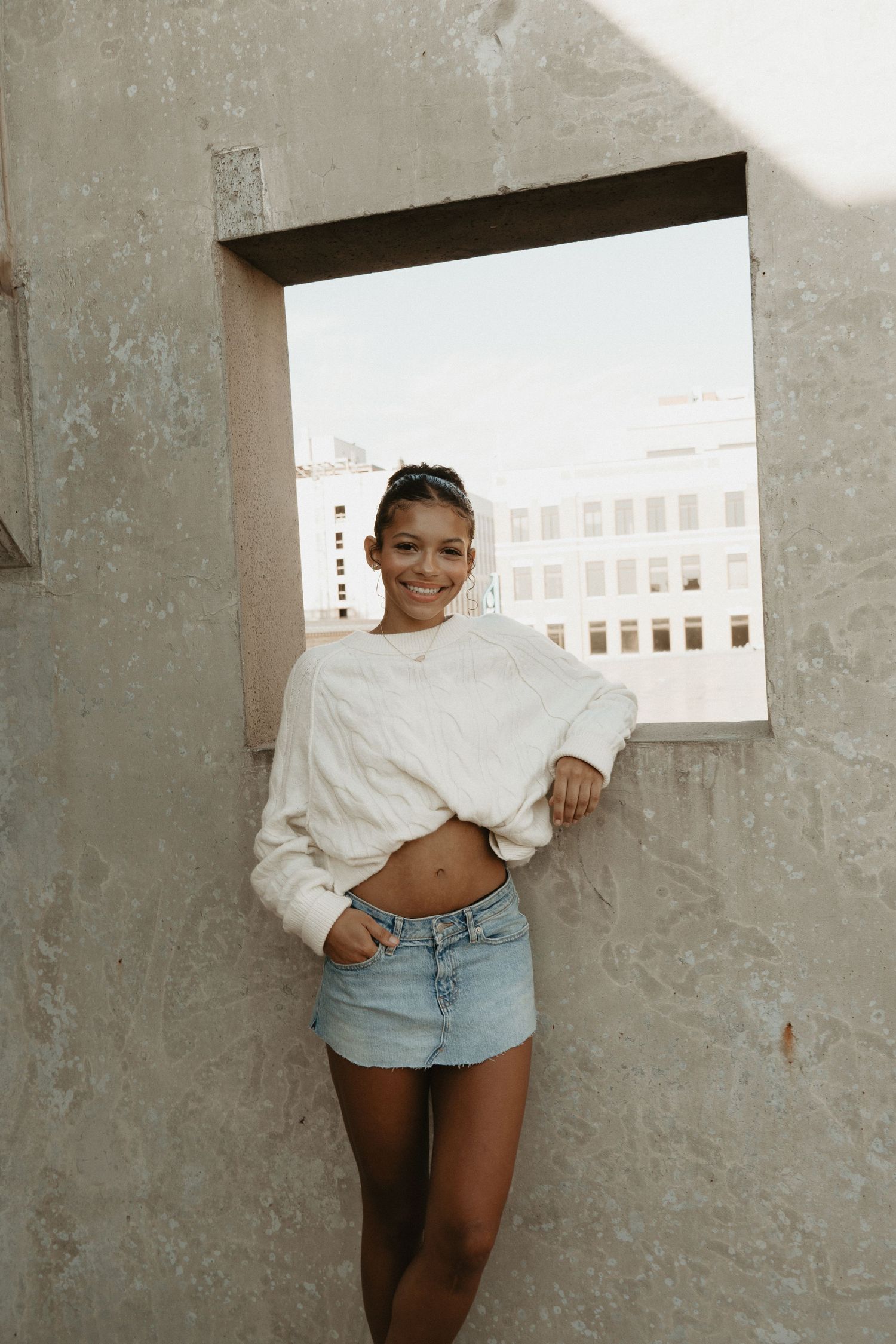 A smiling young woman in a white cropped sweater and denim mini skirt poses on a rooftop.