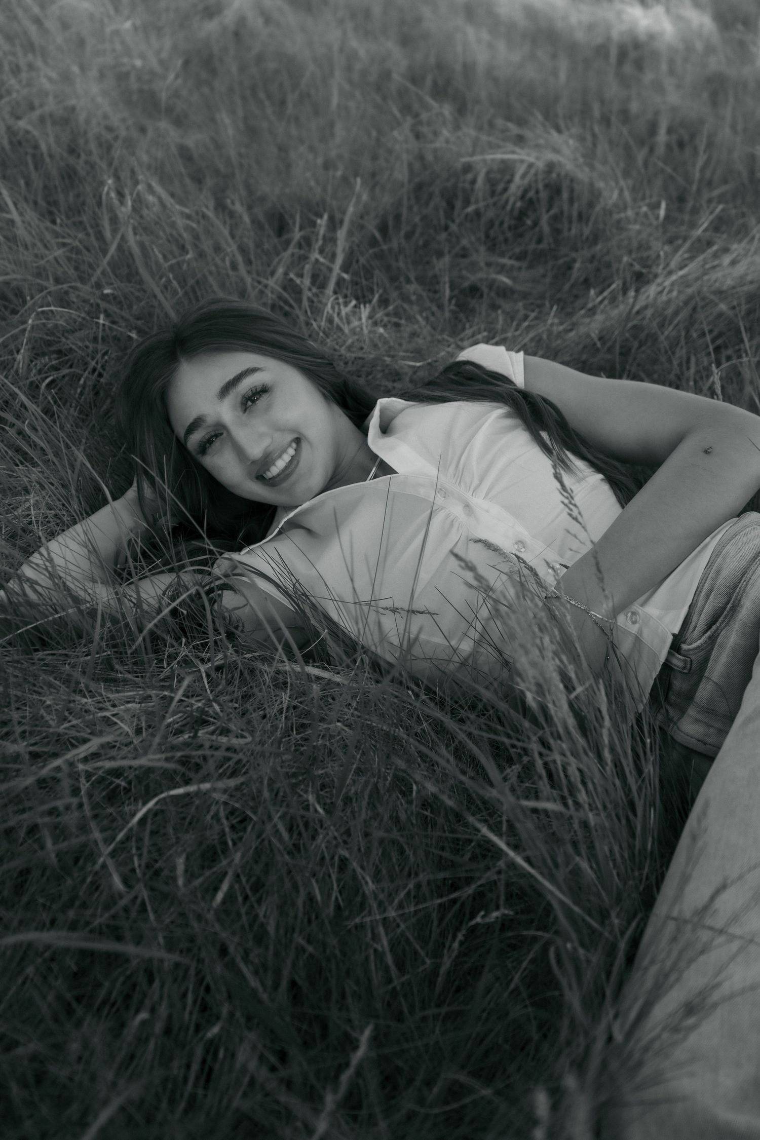 Black and white photo of smiling young woman lying in tall grass field looking up at camera.