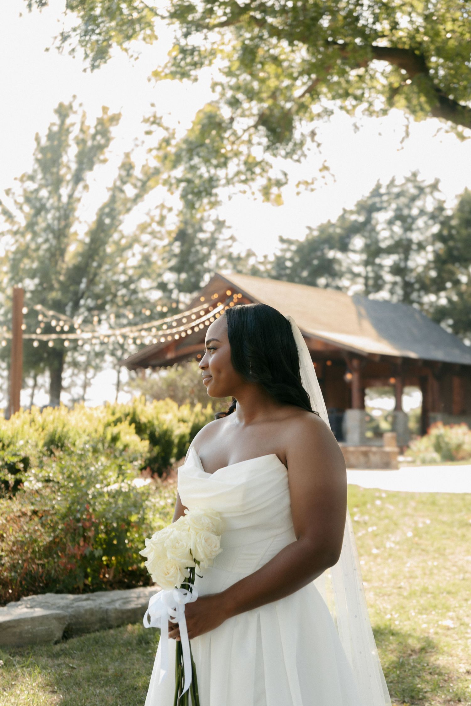 Elegant bride in strapless white gown holding white bouquet standing in sunlit garden at wedding venue.