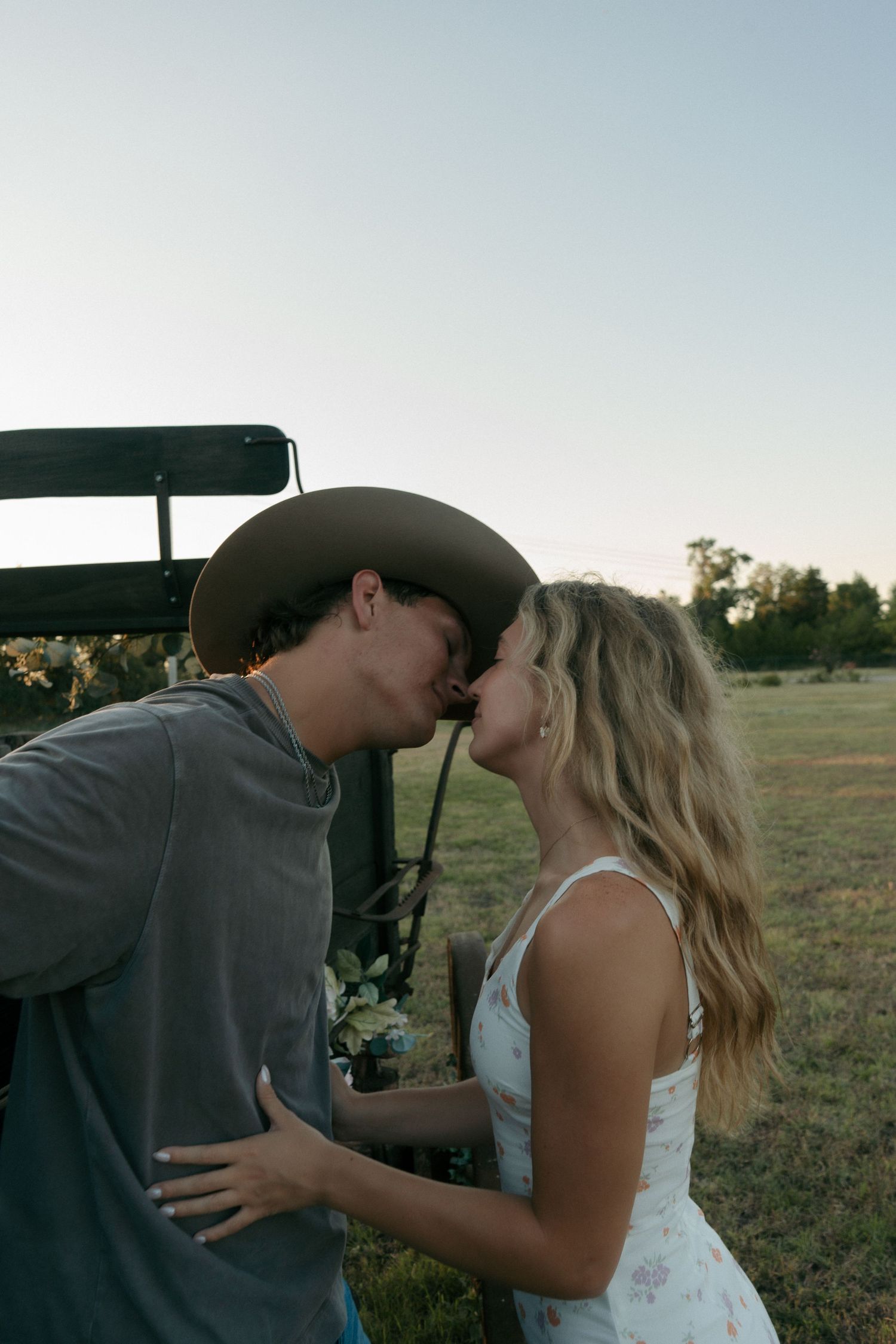 Romantic couple sharing a kiss near a vintage wagon in an open field at dusk.