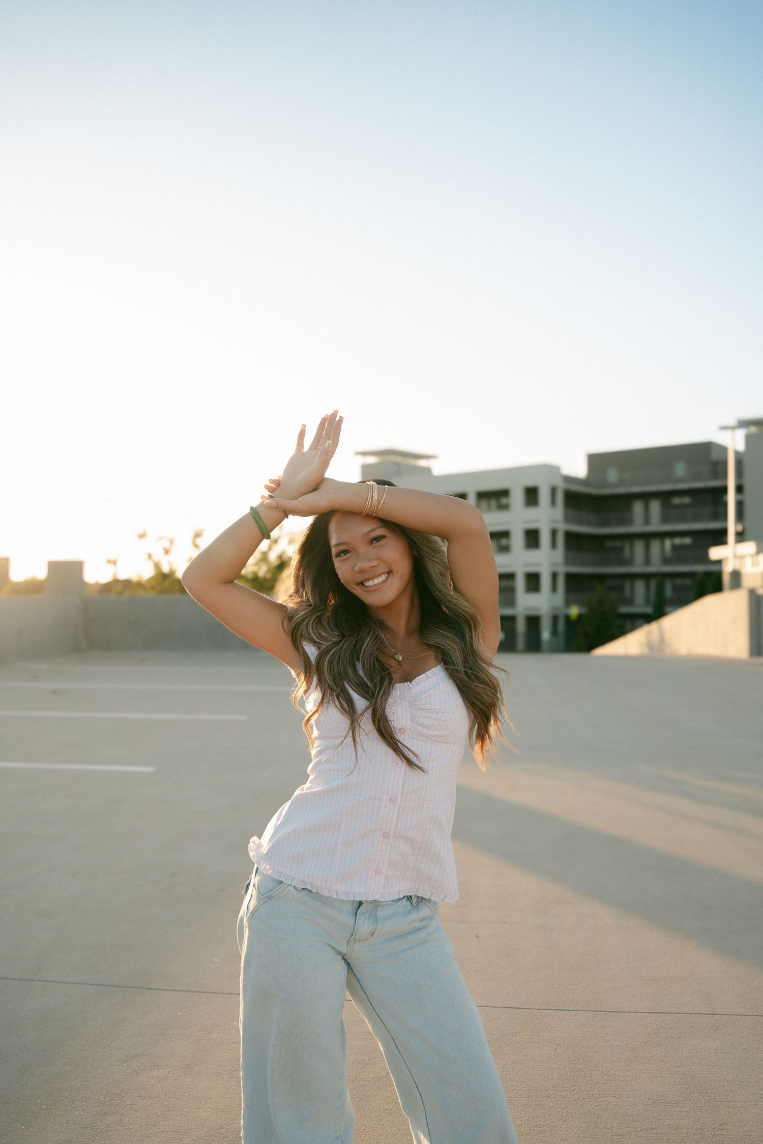 Young woman in white tank top and jeans posing with hand raised at sunny rooftop parking lot.