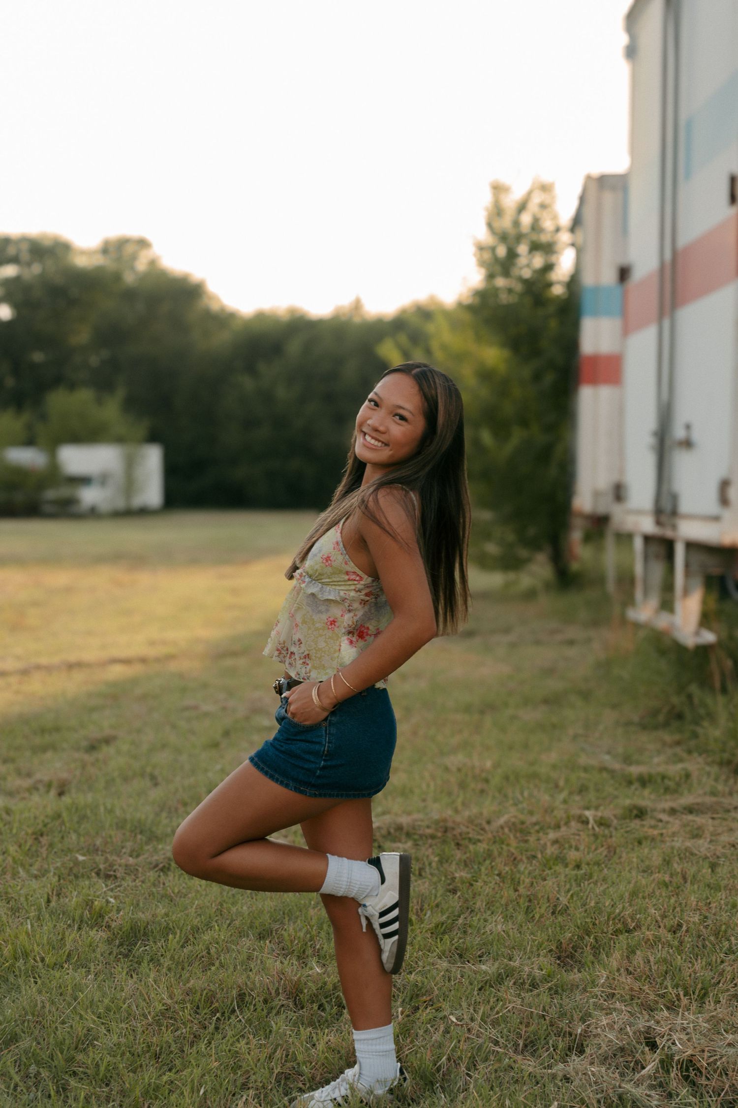 Young woman in floral top and denim skirt posing playfully near a trailer in golden hour light.