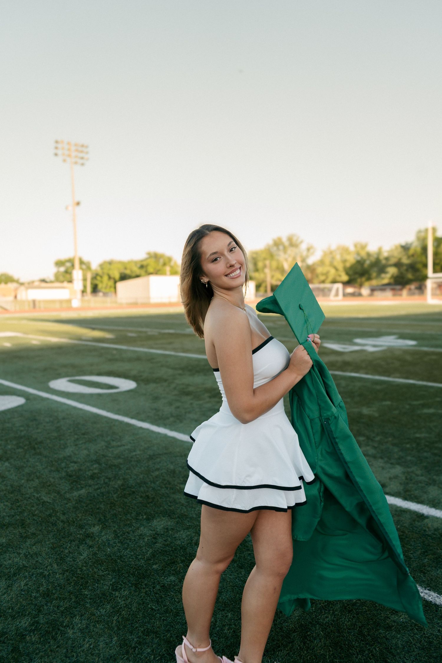 Young woman in white dress holding green graduation cap and gown on field.