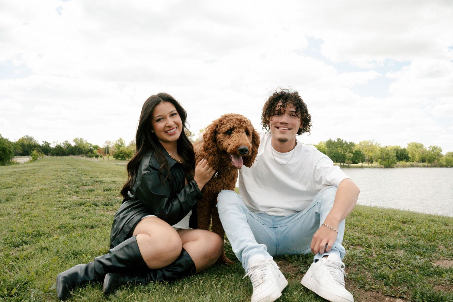 Happy young couple sitting on grass by a lake with their golden doodle dog between them.