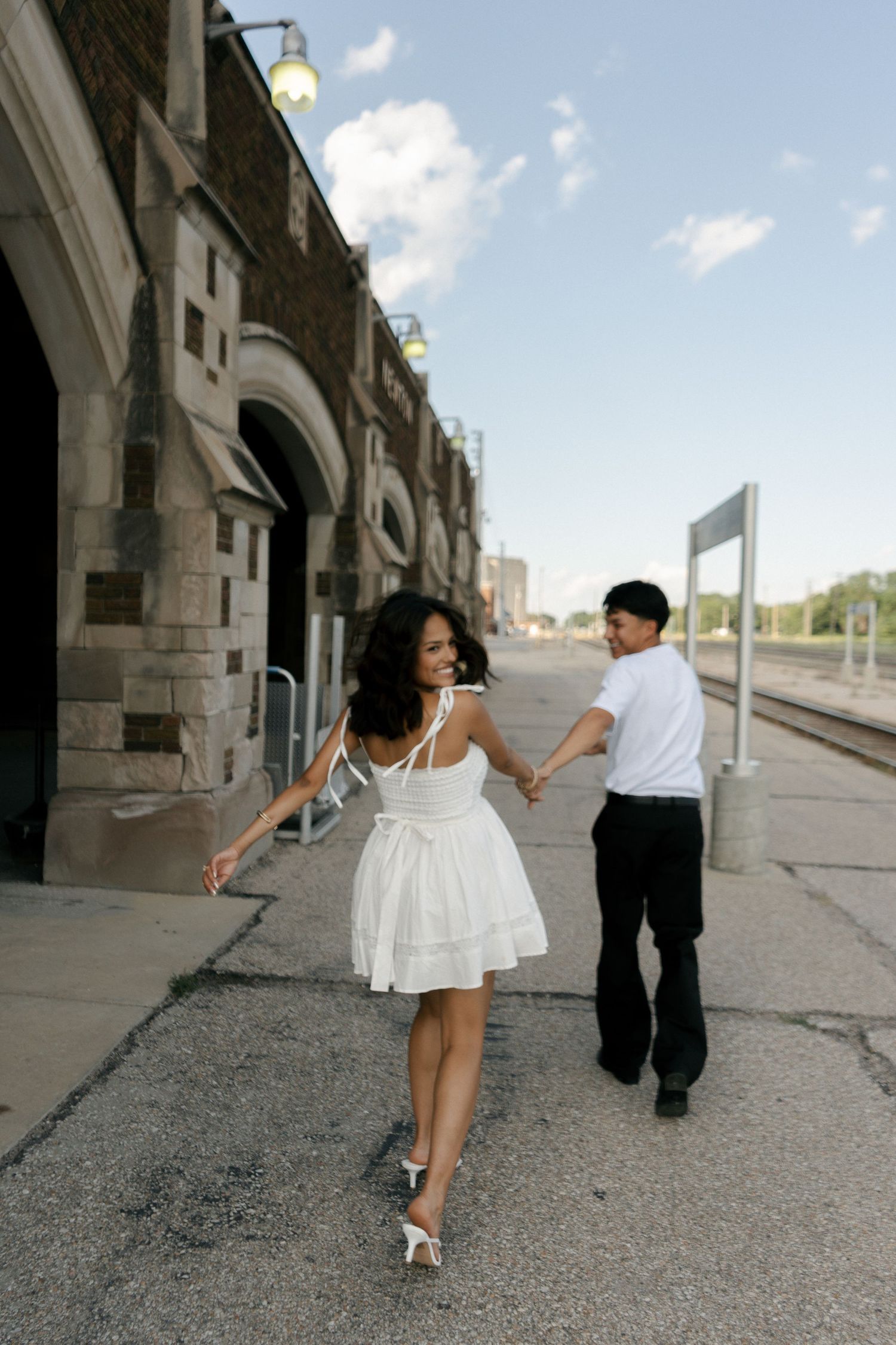 A couple holds hands and dances together on a train station platform, woman in white dress and heels.