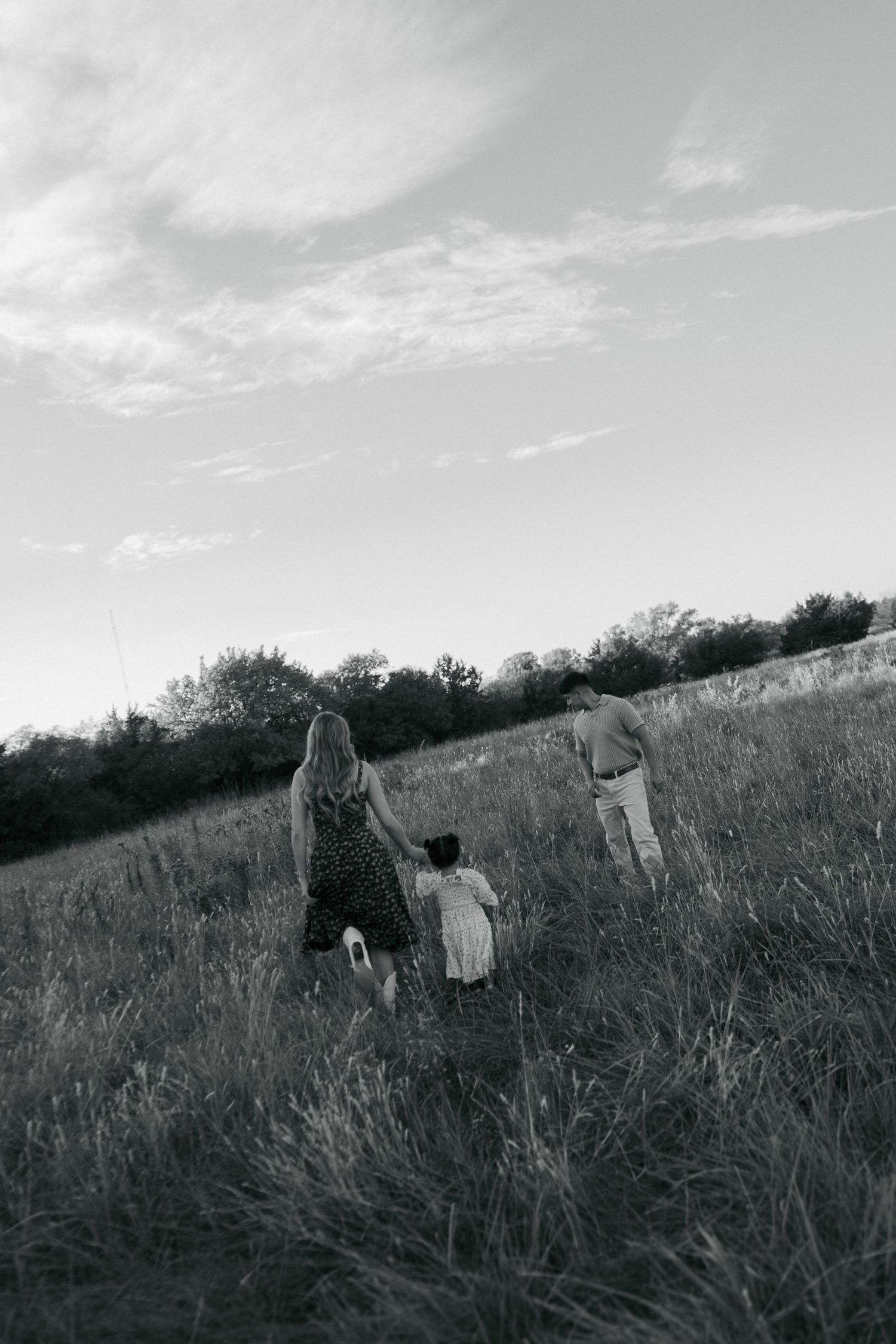 A black and white image of a family walking together through tall grass in an open field at sunset.