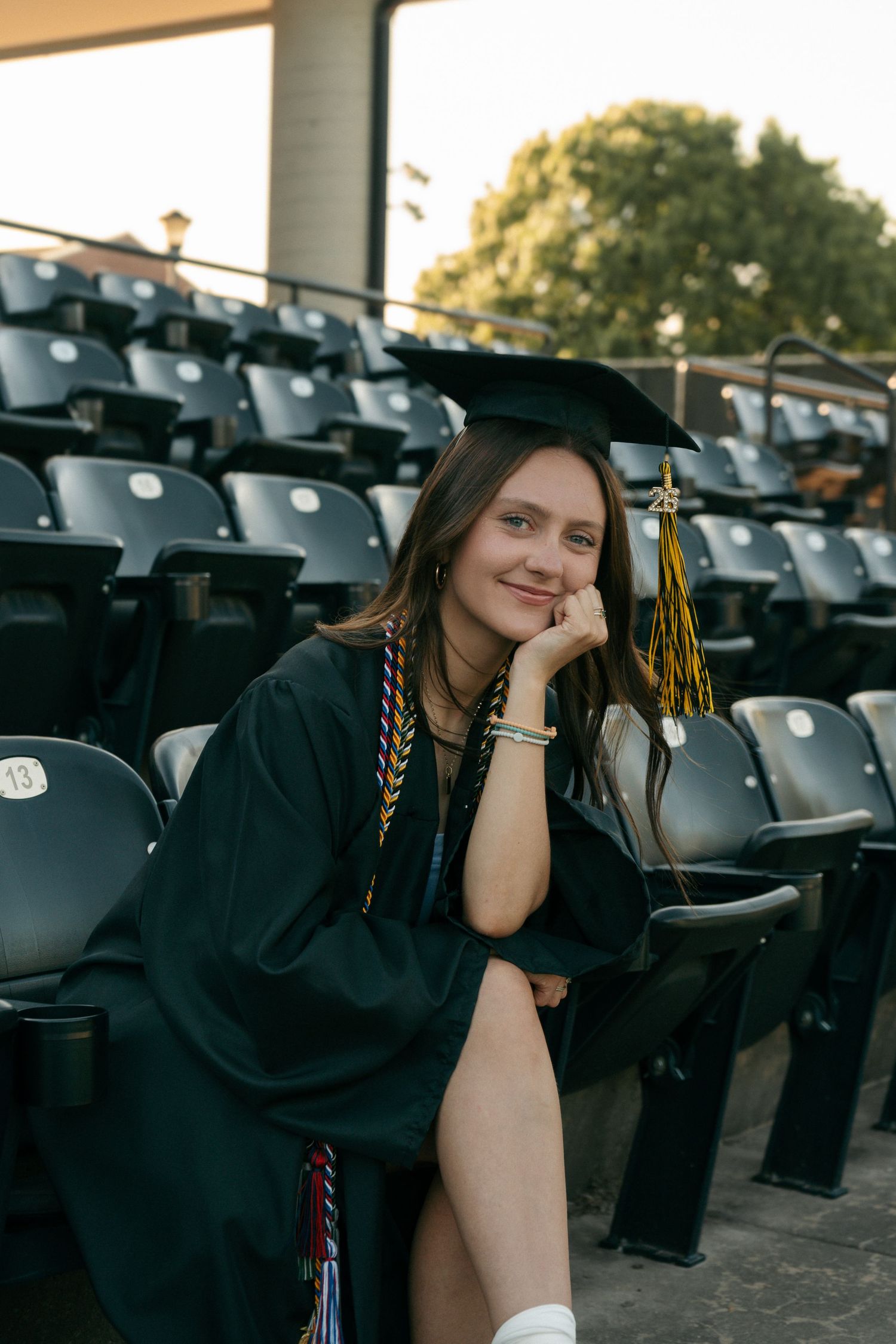 Young woman in green graduation cap and gown smiling while seated in stadium bleachers.