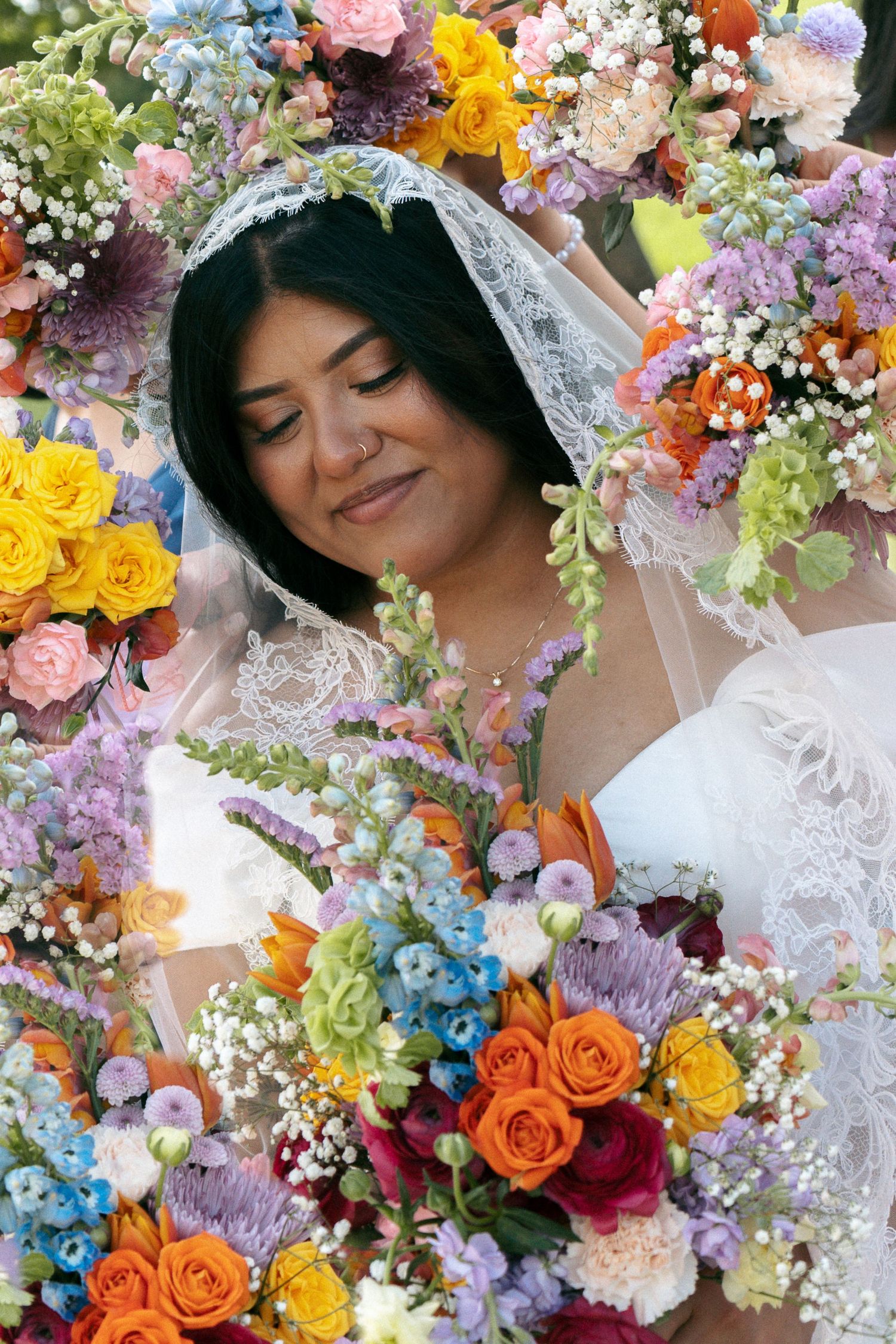Smiling bride in white lace veil surrounded by vibrant colorful wedding bouquets with roses and wildflowers.