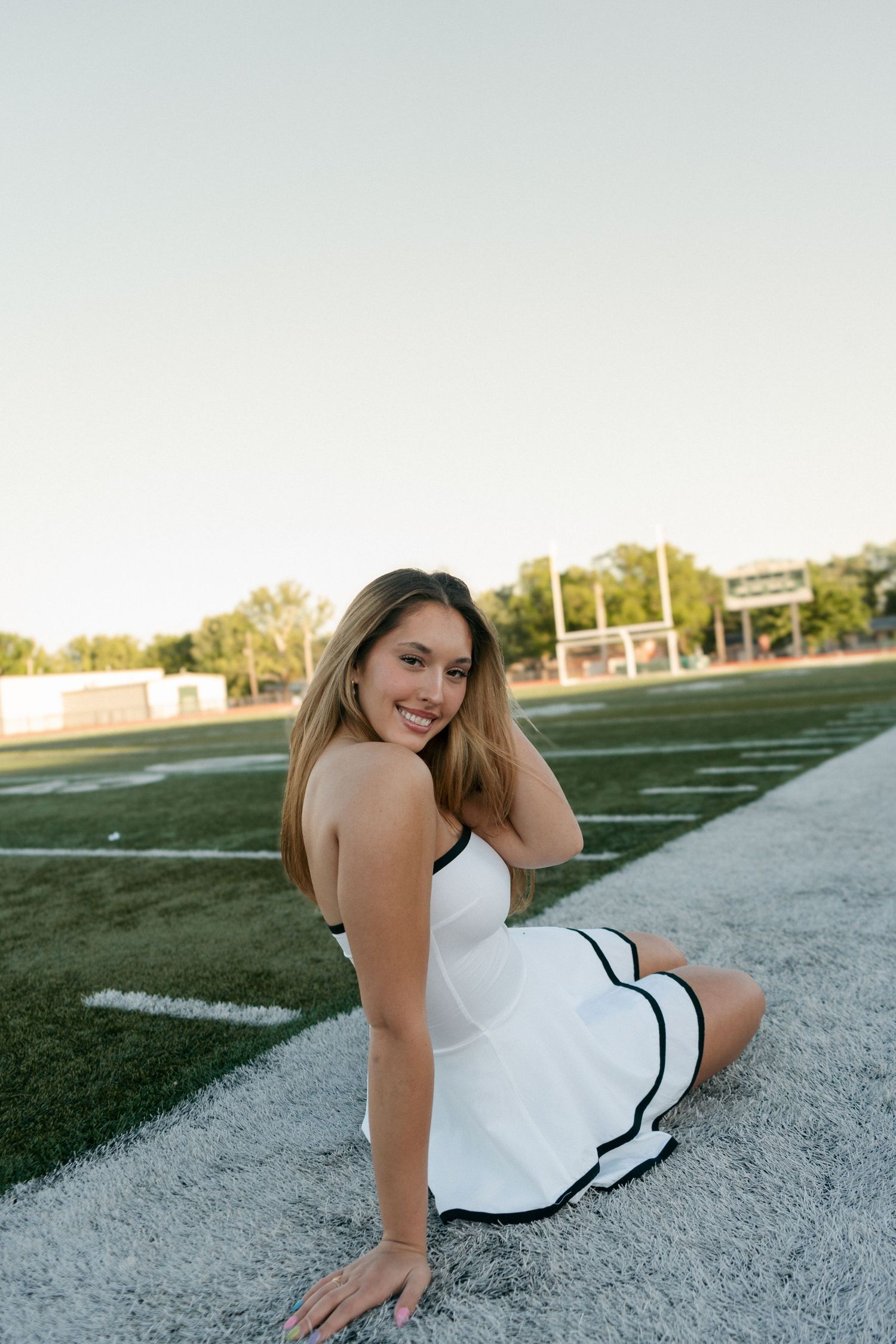 Smiling young woman in white dress with black trim sitting on football field sideline at dusk.