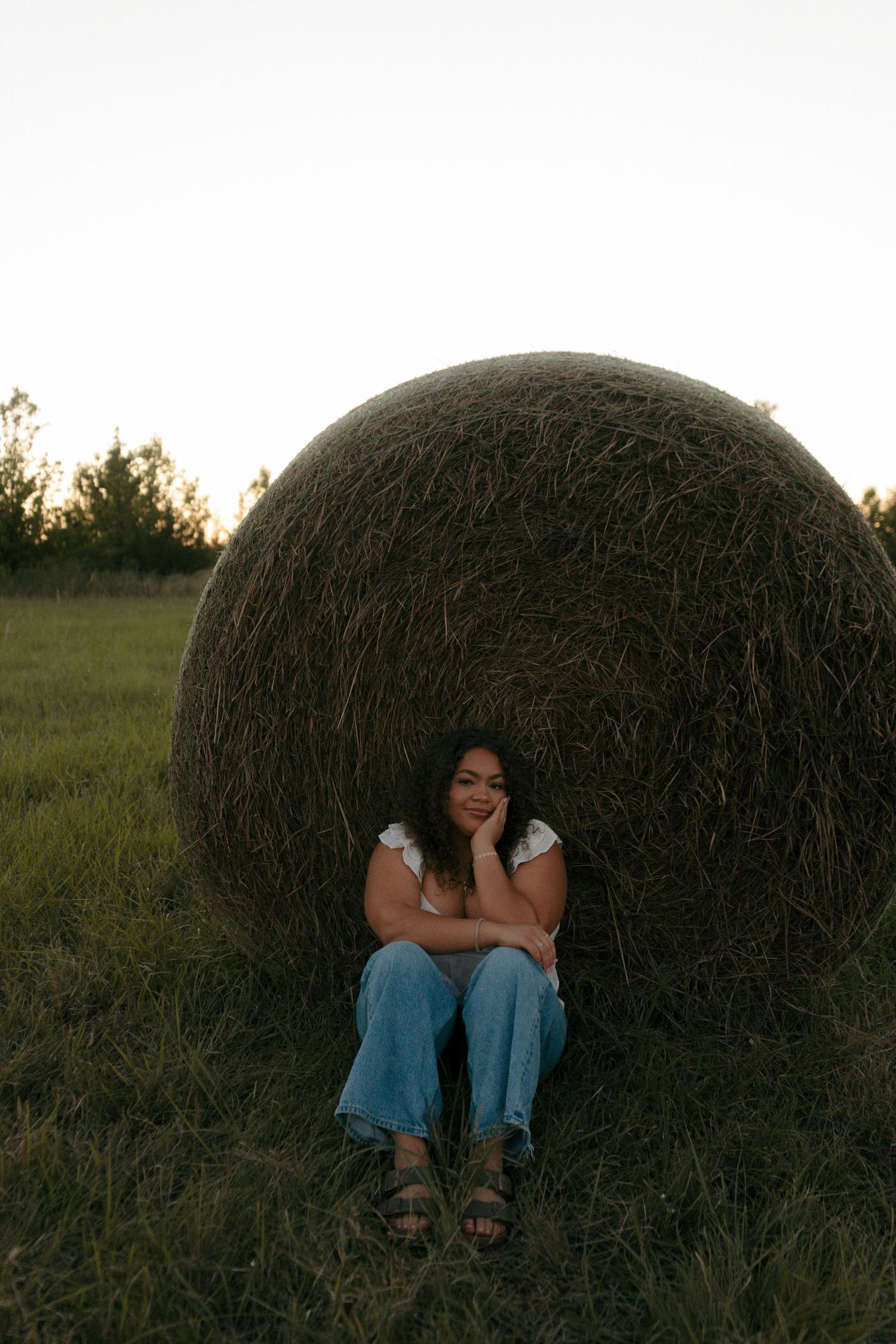 Young woman in white top and jeans sitting against a large hay bale in a field at dusk.